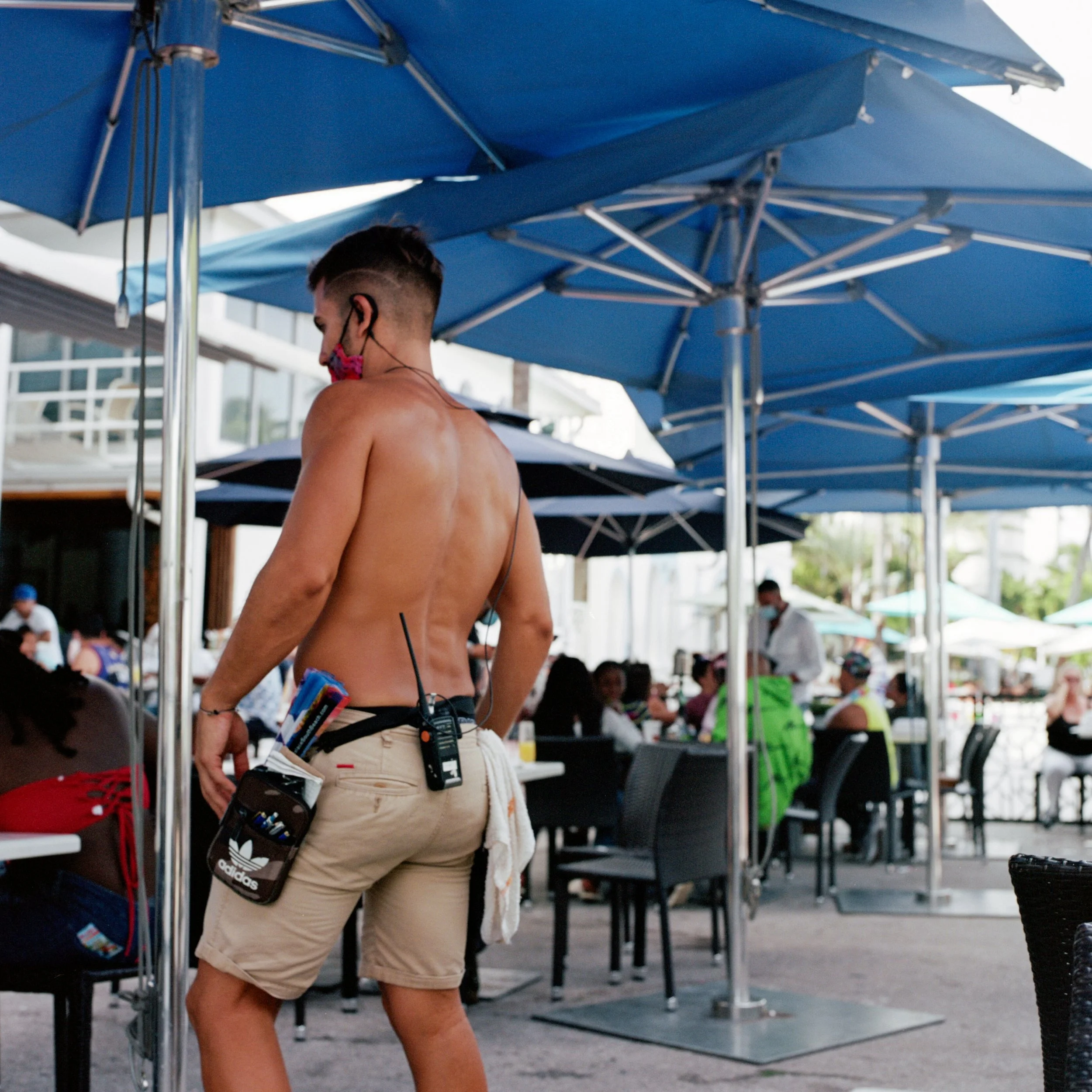 A shirtless man wearing beige shorts and a pink face mask stands under large blue umbrellas at an outdoor dining area with many people seated at tables.