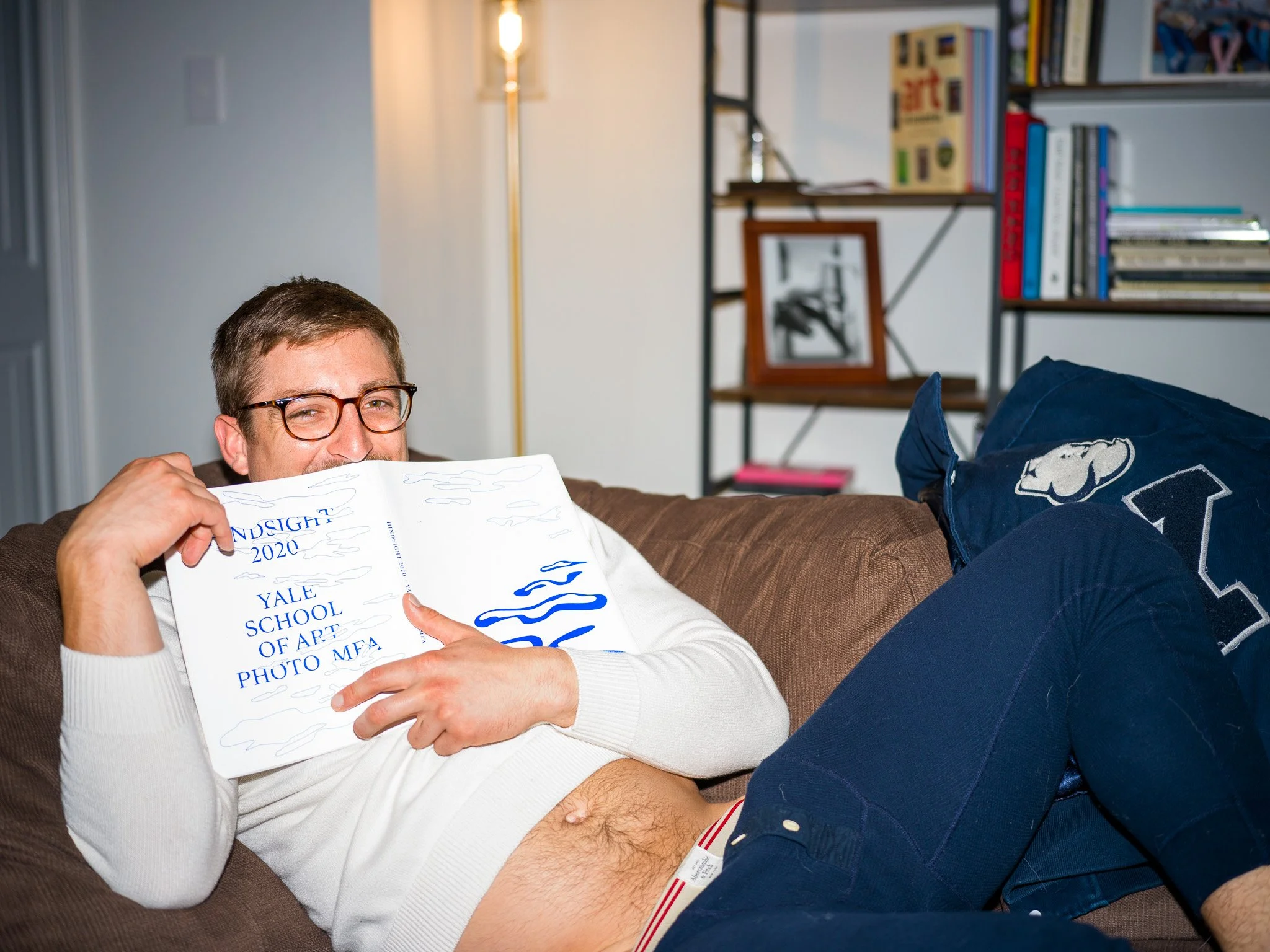 A man with glasses lying on a brown couch, holding an academic pamphlet and smiling. He's partially undressed, with his shirt lifted to reveal part of his abdomen. Bookshelves and framed pictures are in the background inside a room.