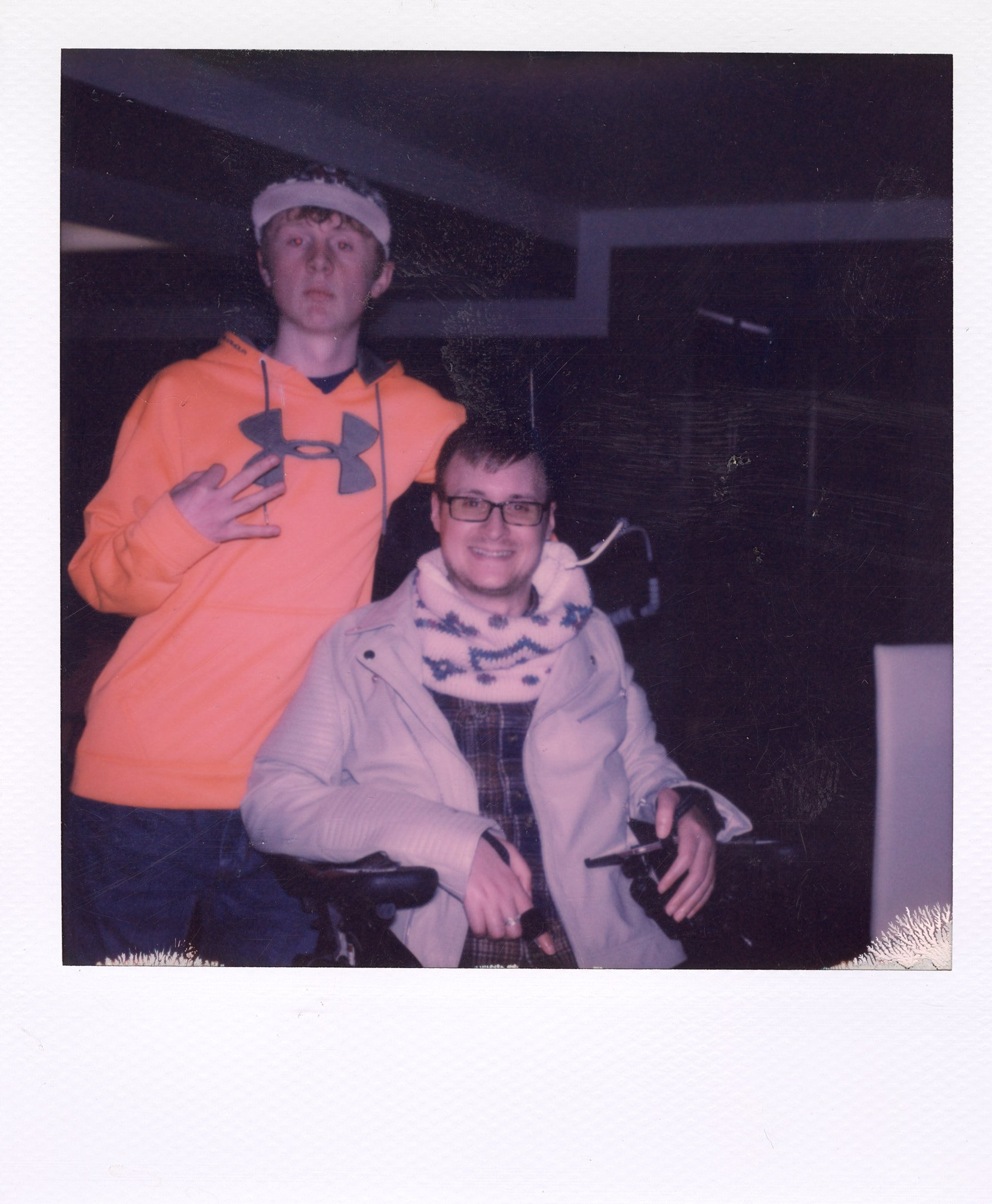 Two young men in an indoor setting, one standing in an orange hoodie making a hand gesture, and the other sitting in a wheelchair wearing glasses, a beige jacket, and a patterned scarf, both smiling at the camera.