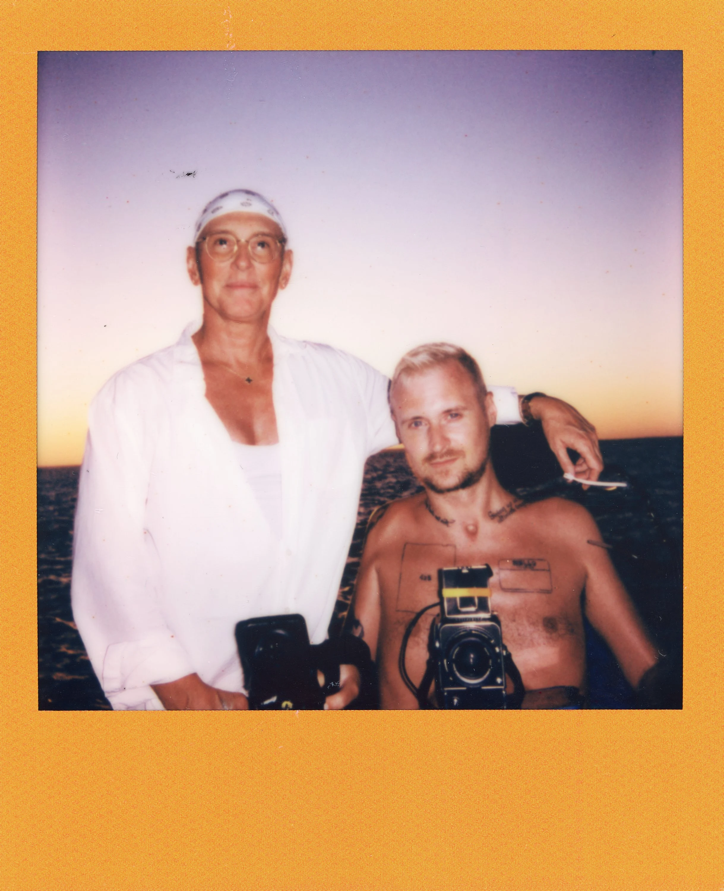 Two men posing together on a boat during sunset, one standing and the other sitting with a camera on his chest.
