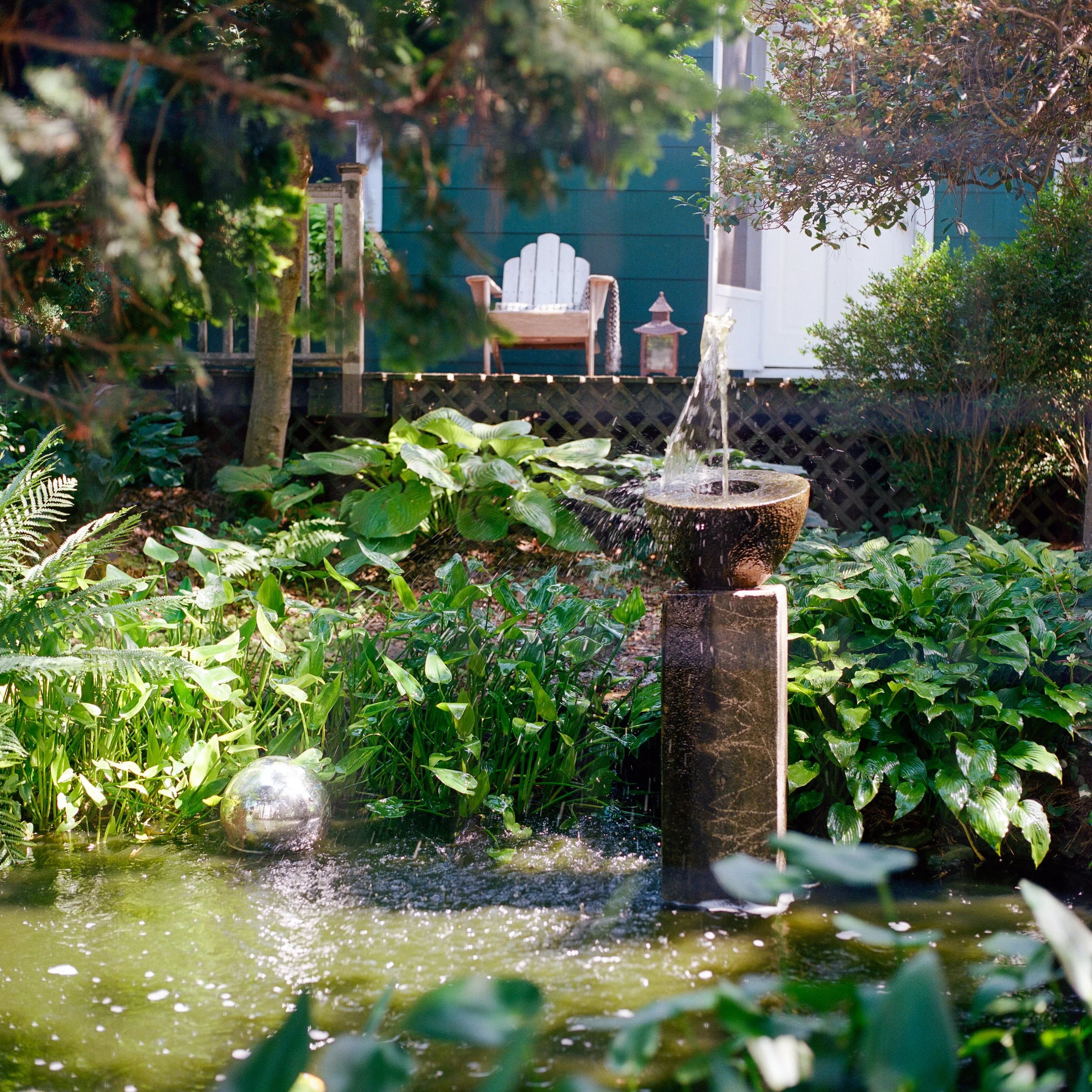 A garden fountain with water flowing into a pond, surrounded by lush green plants and trees, with a wooden chair and small lantern on a deck in the background.