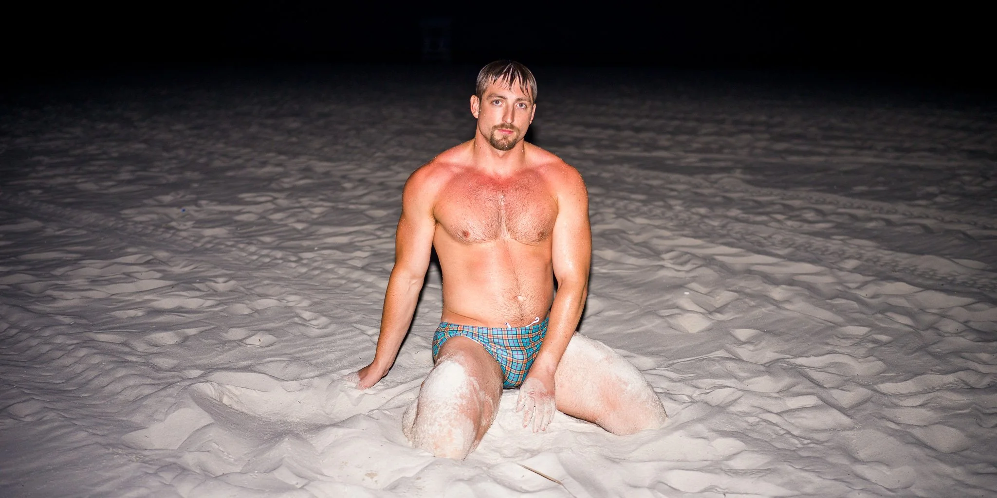 A shirtless man in plaid swim trunks kneels in the sand at night, with a dark sky overhead, looking directly at the camera.