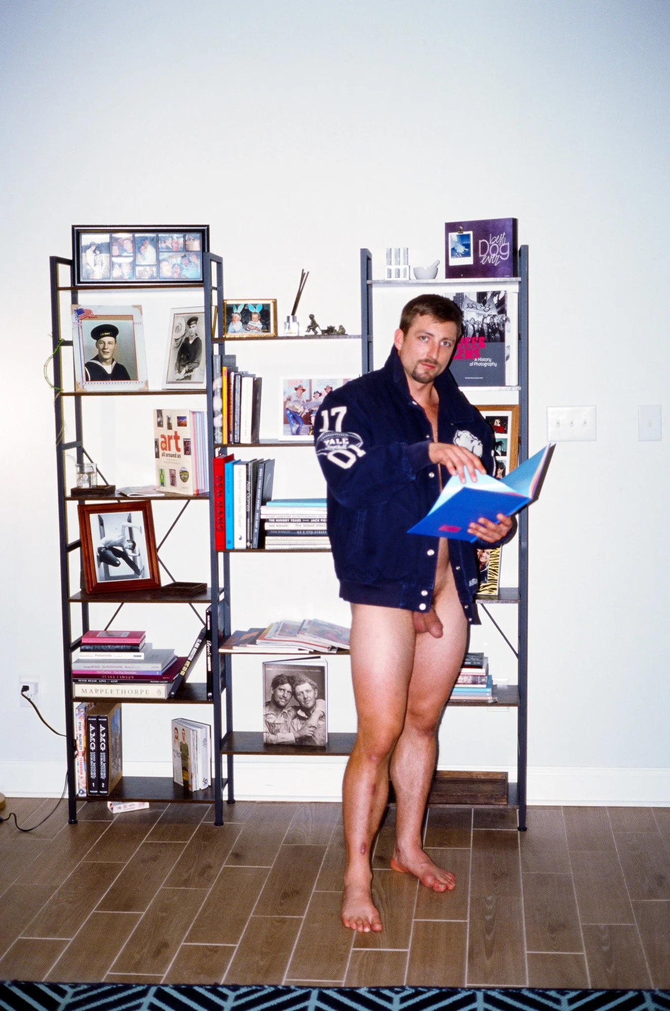 A man standing indoors, wearing an open jacket, holding a book, and with no pants, revealing his genital area. He is barefoot in front of a bookshelf filled with various books and framed photos.