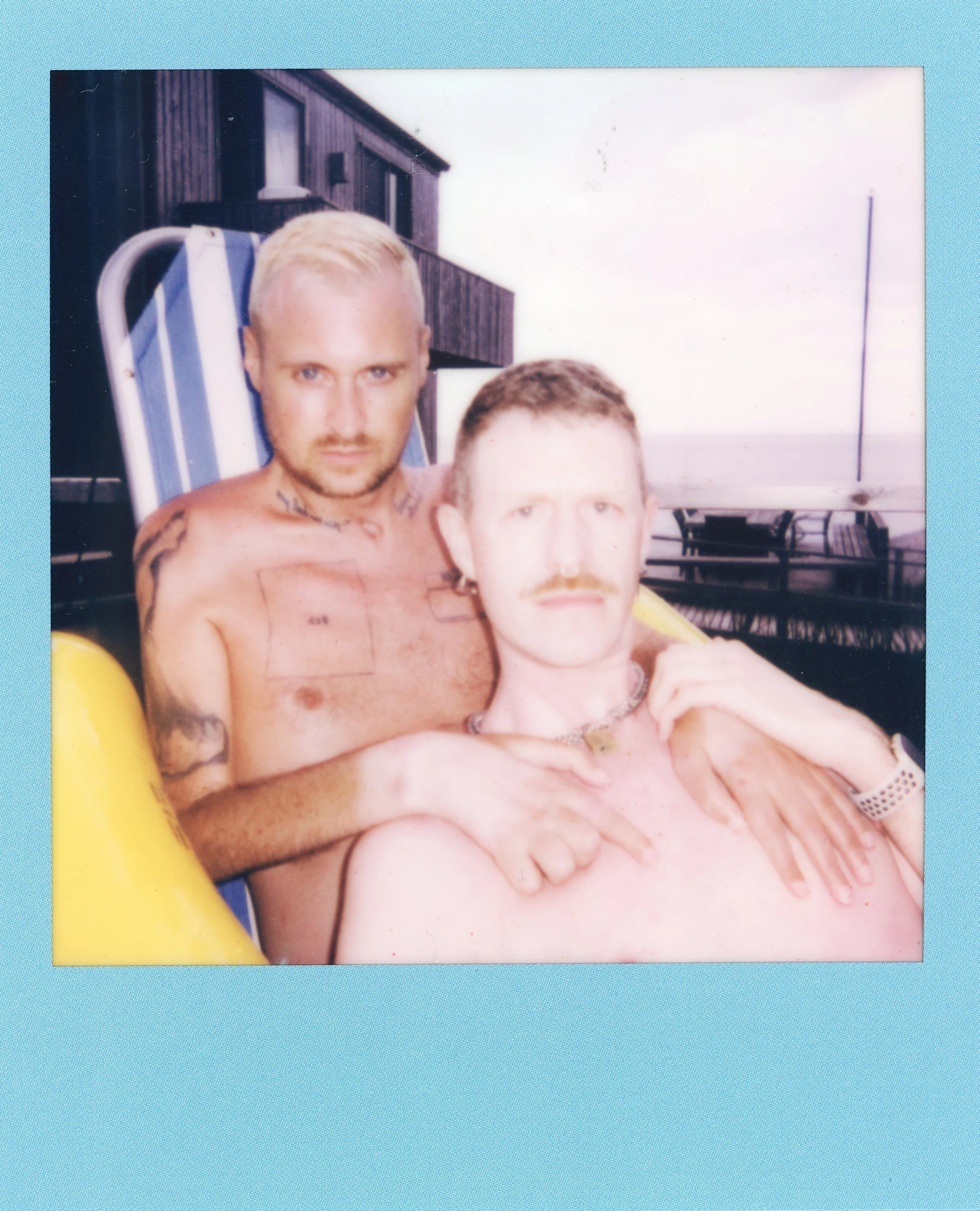 Two shirtless men relaxing outdoors on a deck or balcony, with a building and water in the background, one with tattoos and the other with a mustache and wearing a watch and necklace.