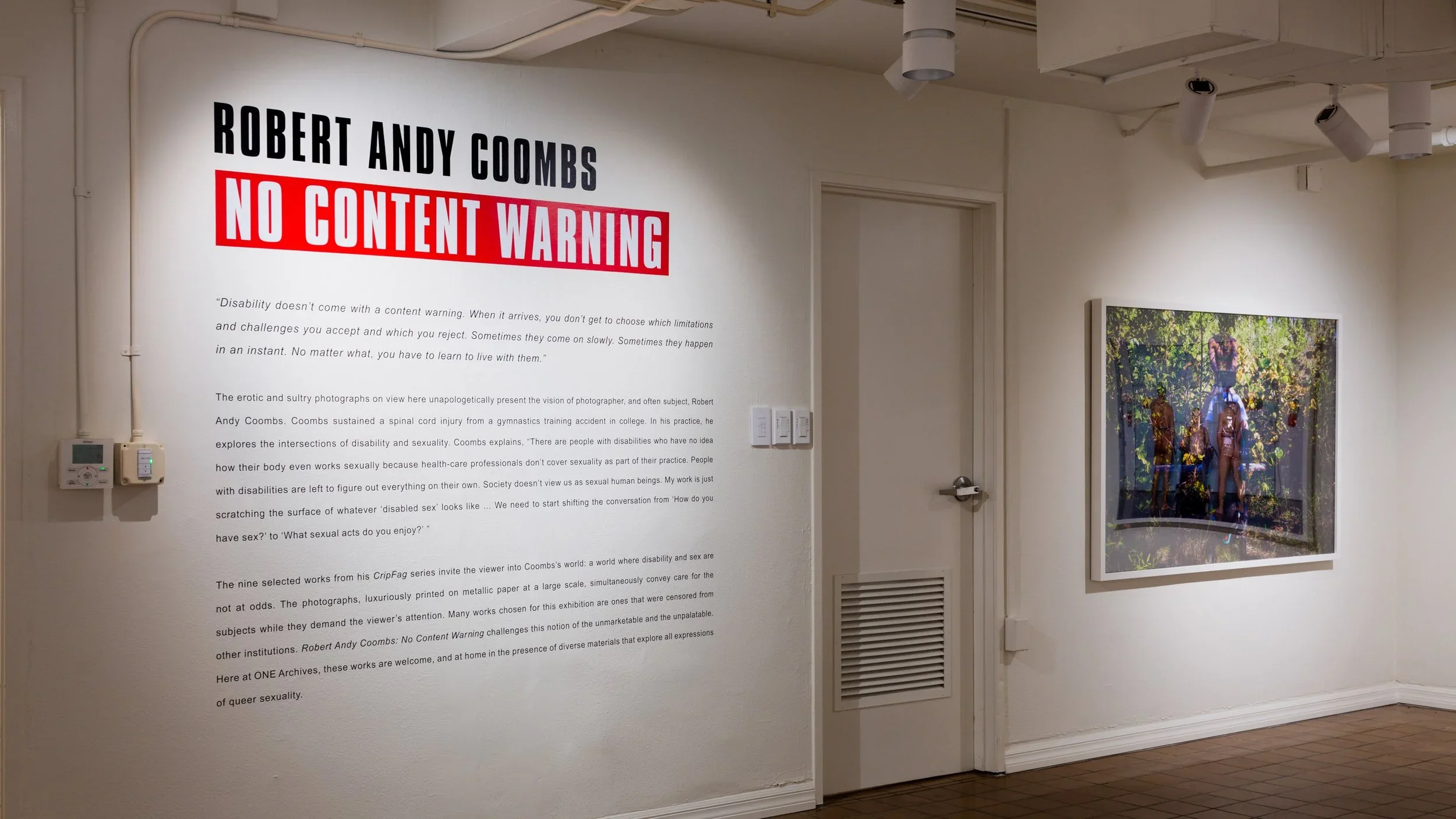 An art gallery wall features a large text panel titled 'ROBERT ANDY COOMBS NO CONTENT WARNING' with paragraphs of description underneath. To the right, a framed photograph depicts three human figures, partially transparent, standing outdoors among trees and foliage.