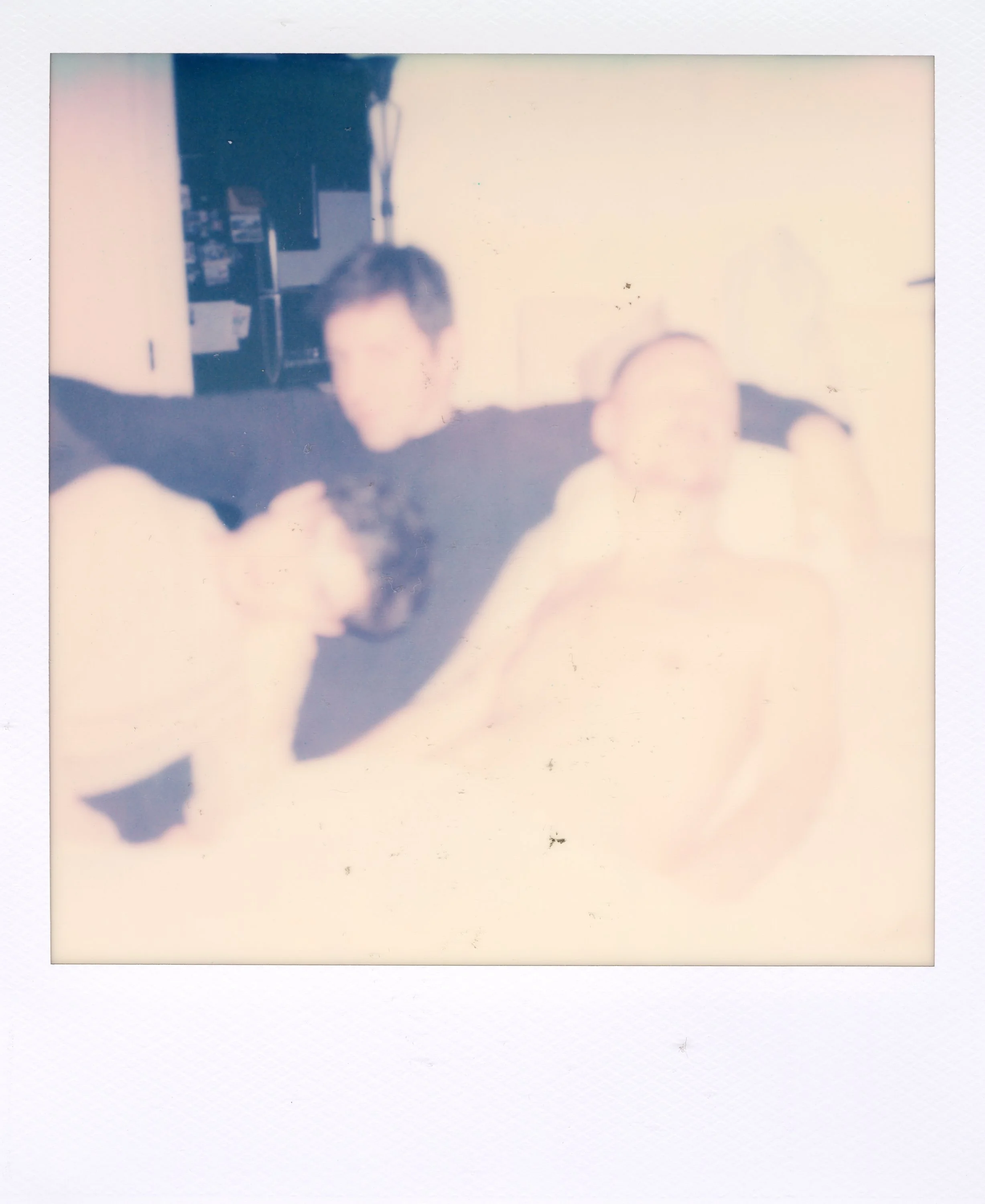 A blurry Polaroid photo of two people sitting on a couch, one with arms outstretched, in a room with a refrigerator in the background.