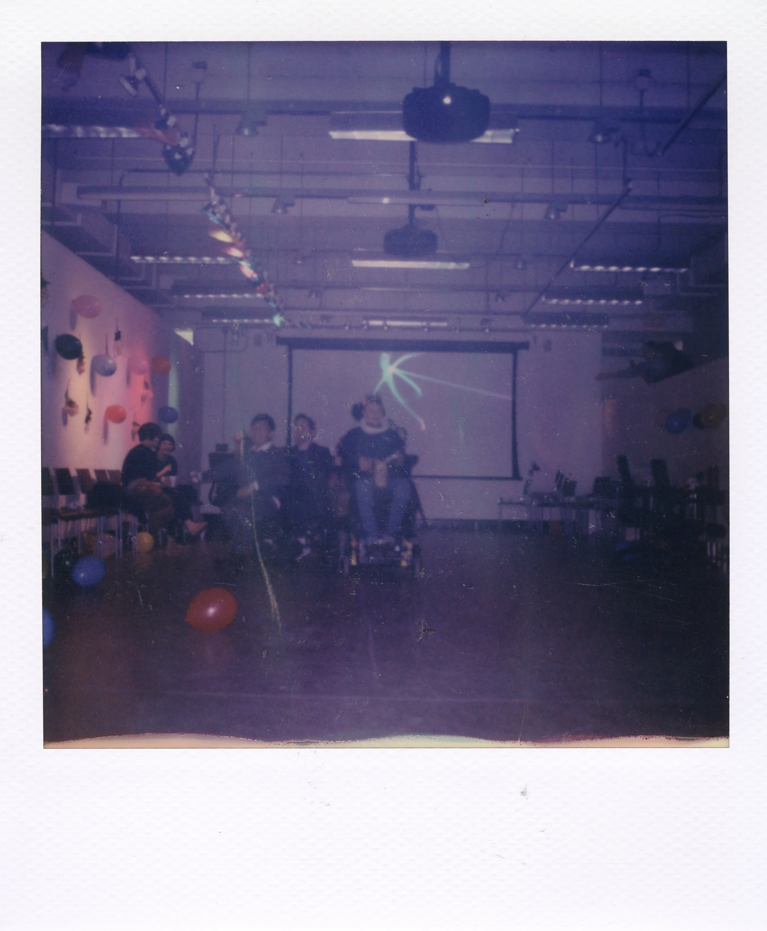 A room decorated with balloons and streamers, with a group of people standing in front of a projection screen and some chairs along the sides.