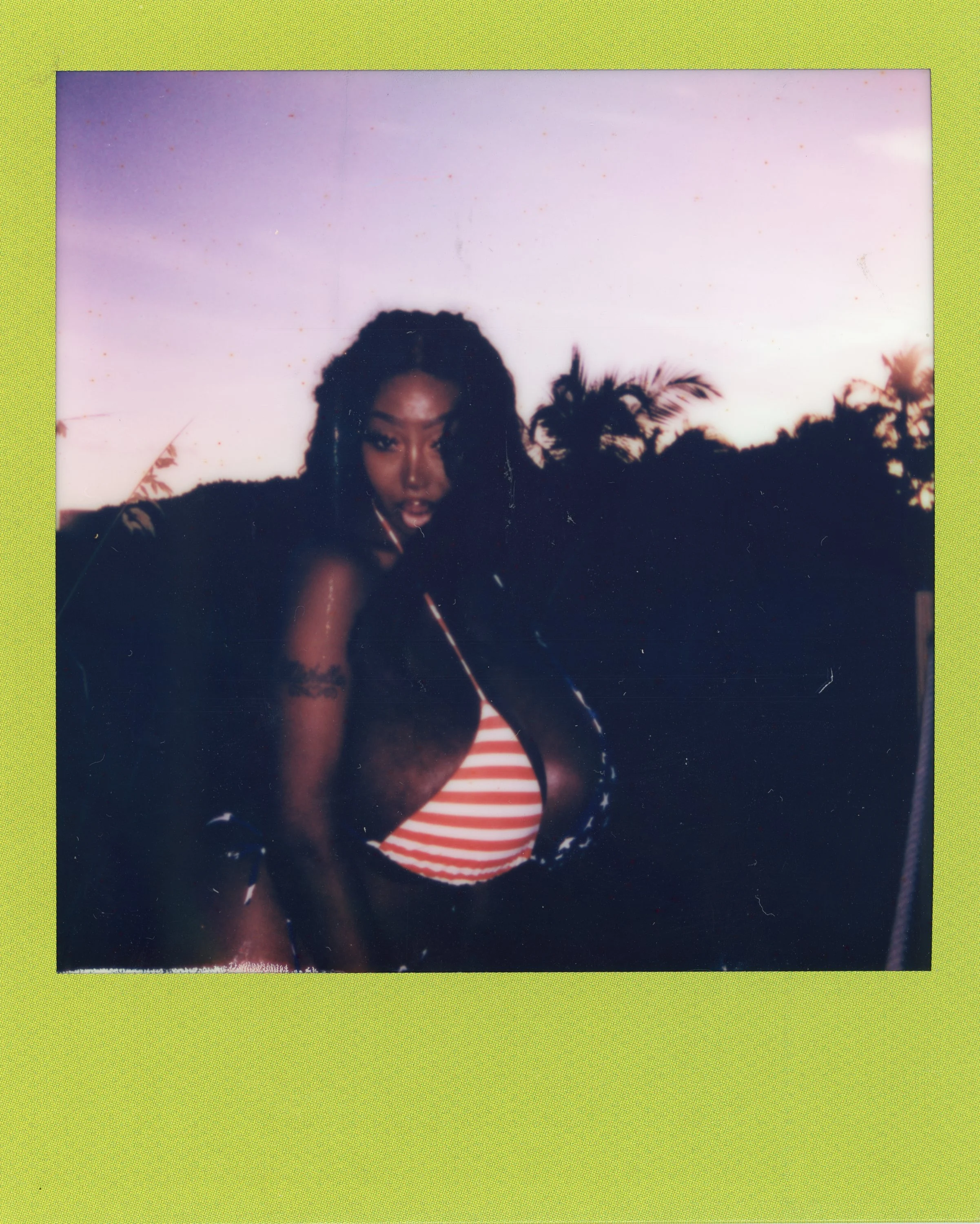 A woman with curly hair posing outdoors during sunset, wearing a striped top with a black cover-up, against a backdrop of palm trees.