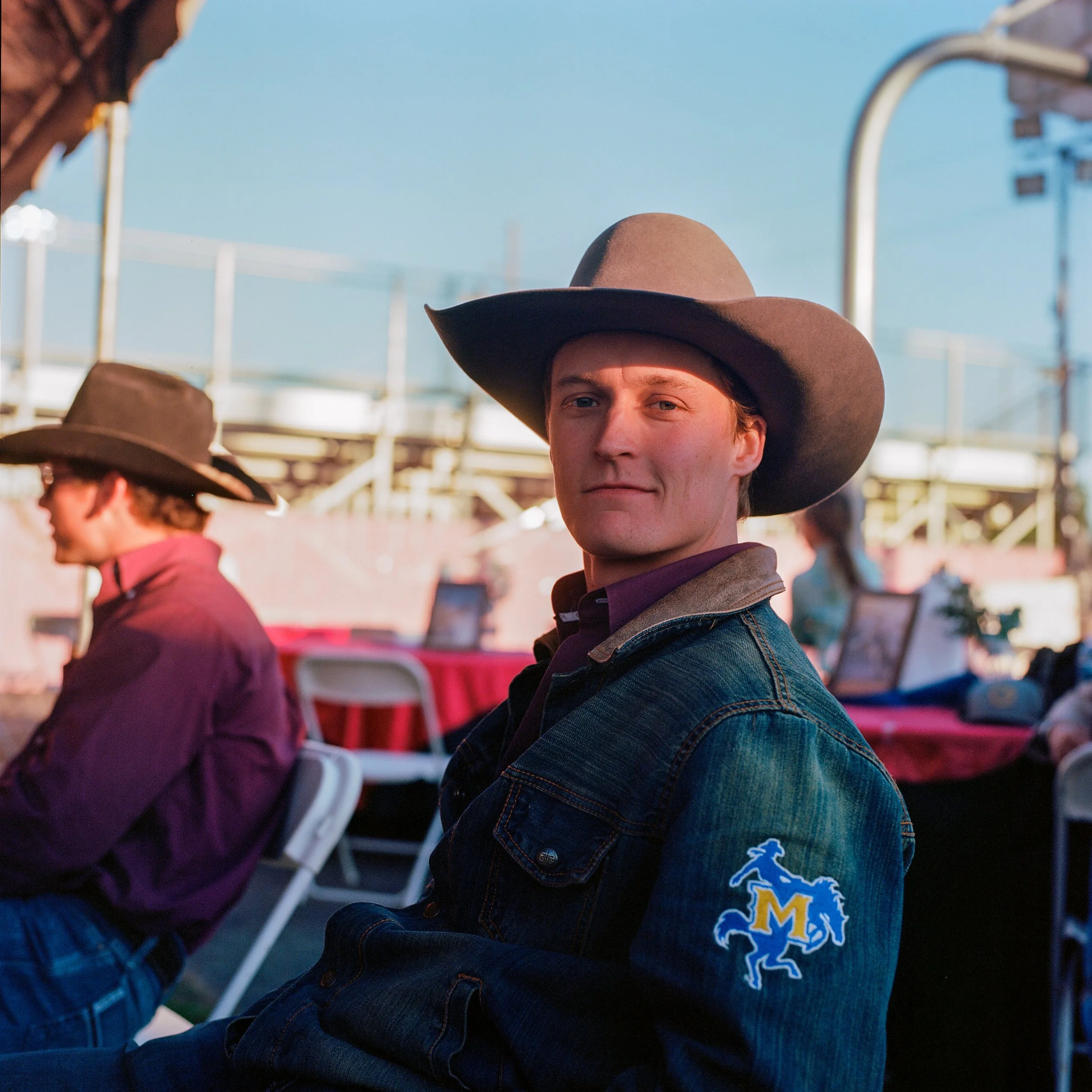 A young man wearing a cowboy hat and denim jacket with an emblem sits outdoors at an event with other people in the background.
