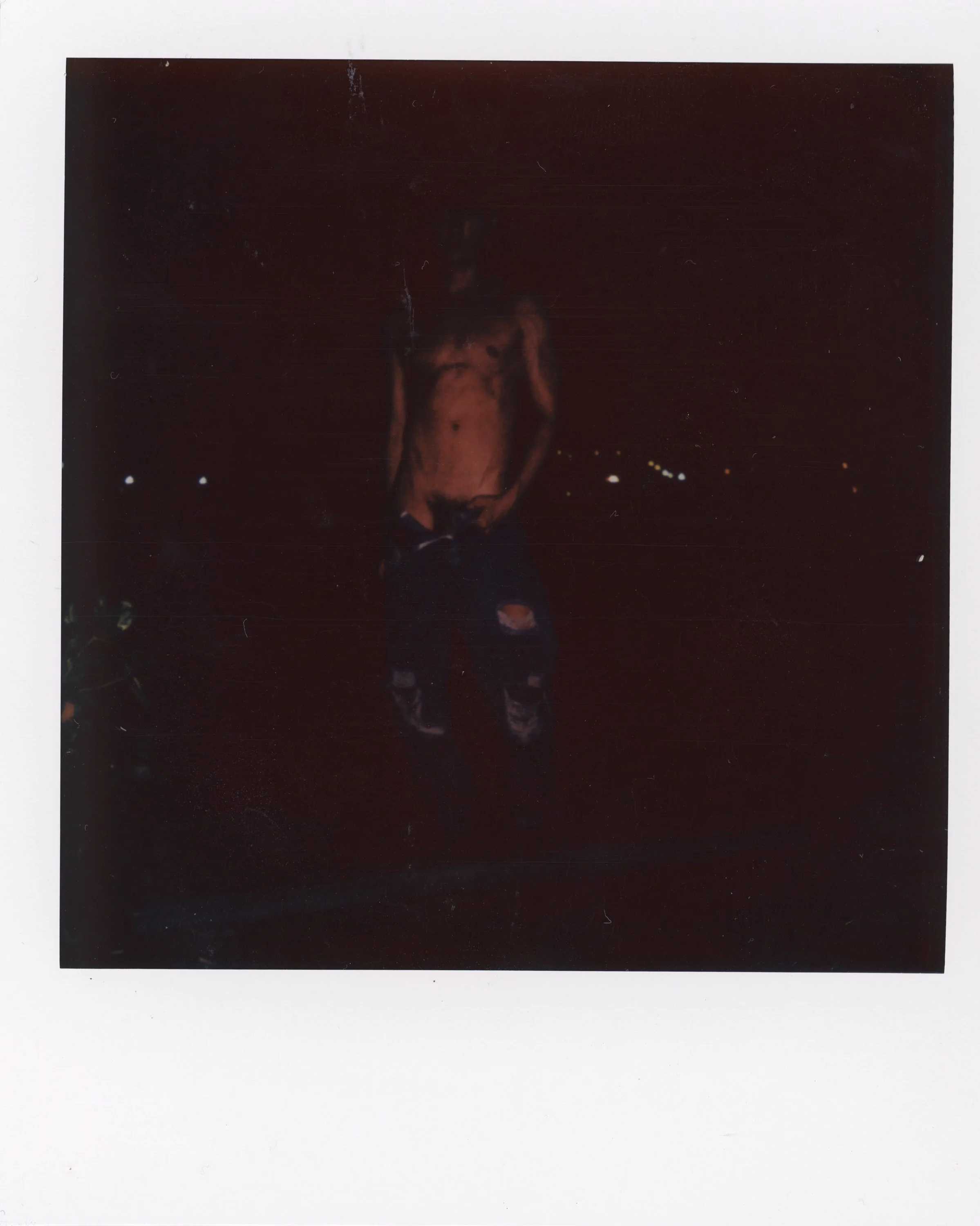 A shirtless man with ripped jeans standing outdoors at night. He is holding something in his right hand, and there are small lights in the background.