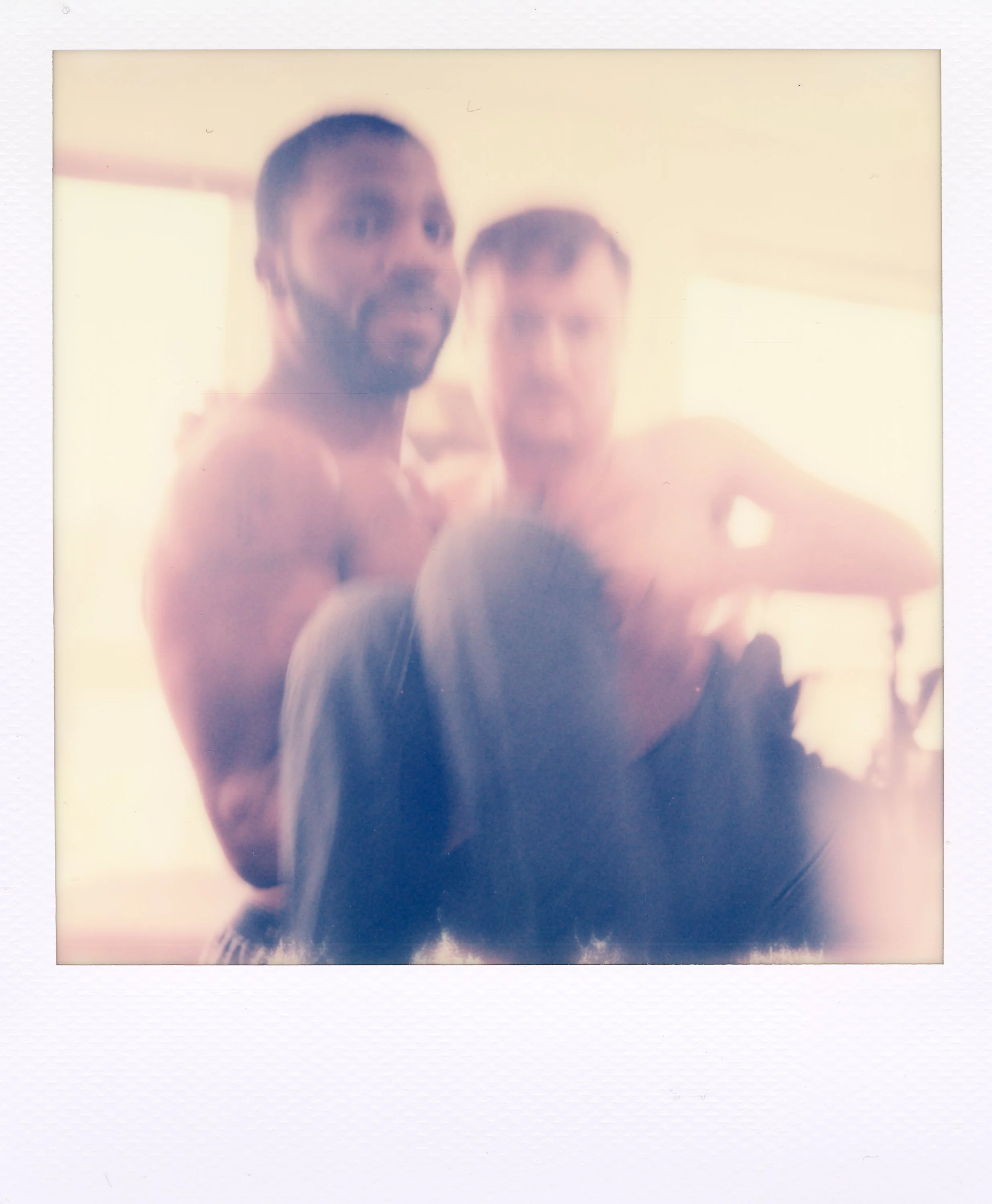 Two shirtless men with a woman in black clothing are posing for a photo in a bright room with natural light.