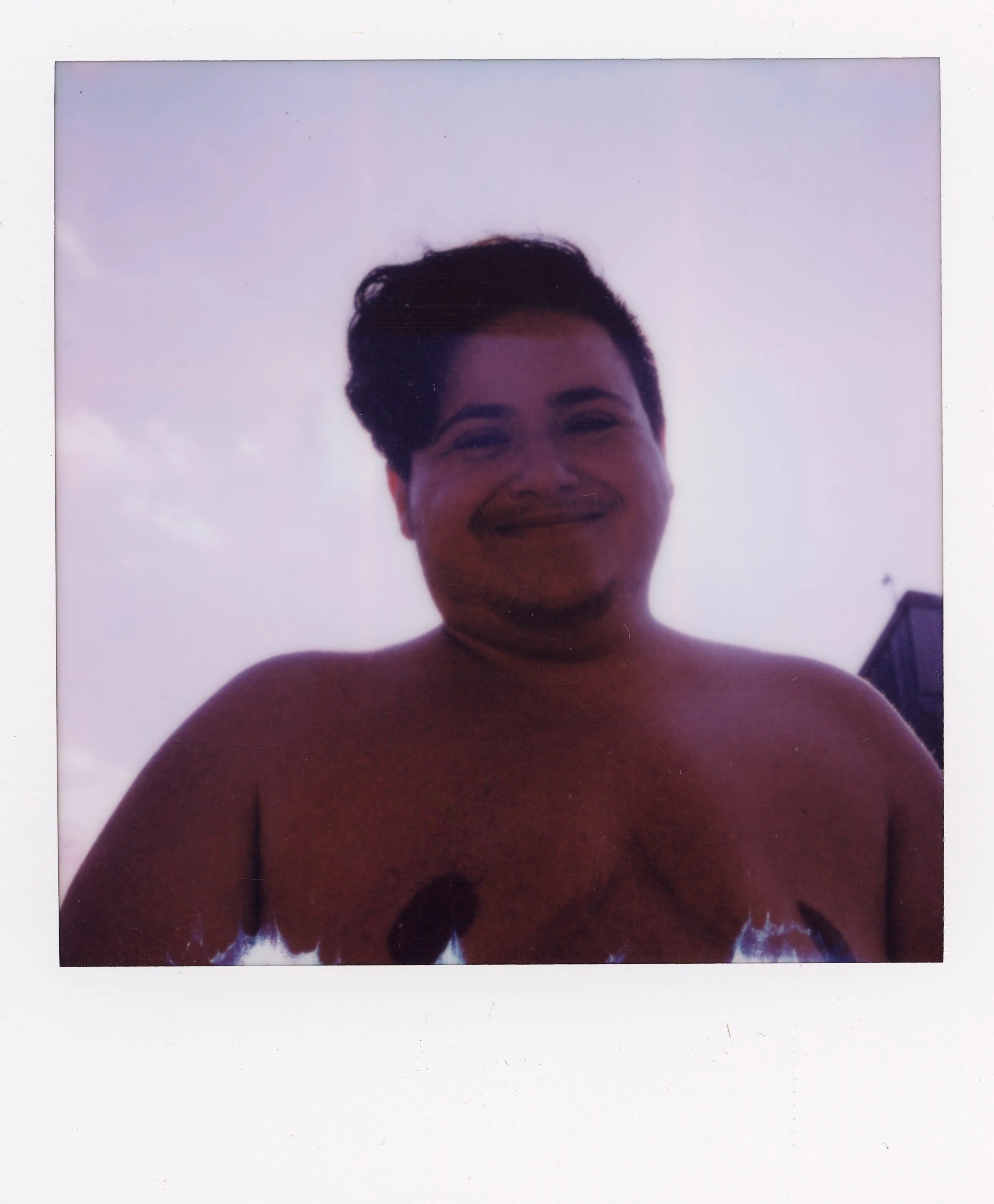 A smiling person with short dark hair and no shirt stands outdoors against a cloudy sky and a building in the background.