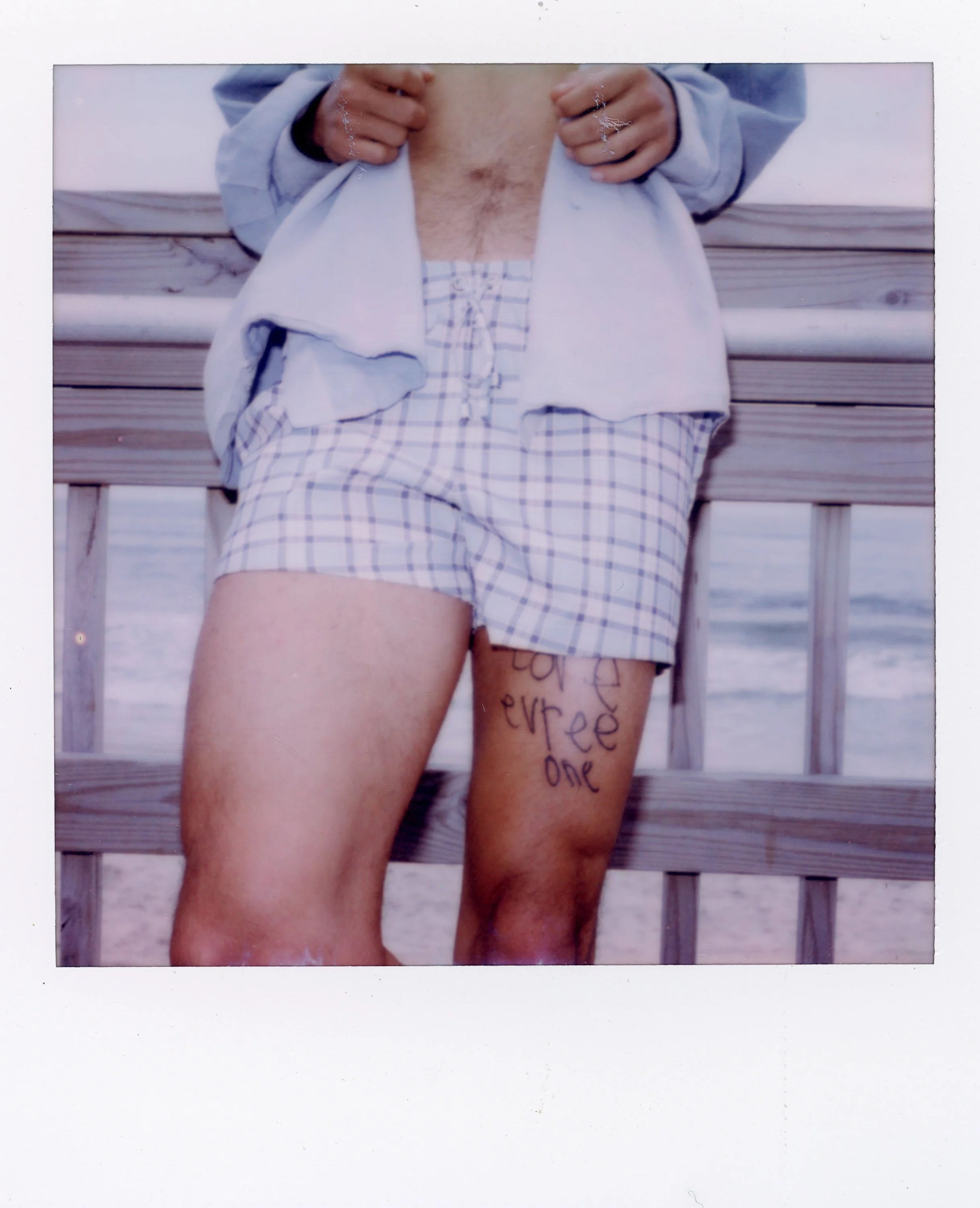 Person standing on a beach wearing plaid shorts, lifting a shirt to reveal their stomach, with writing on their thigh.