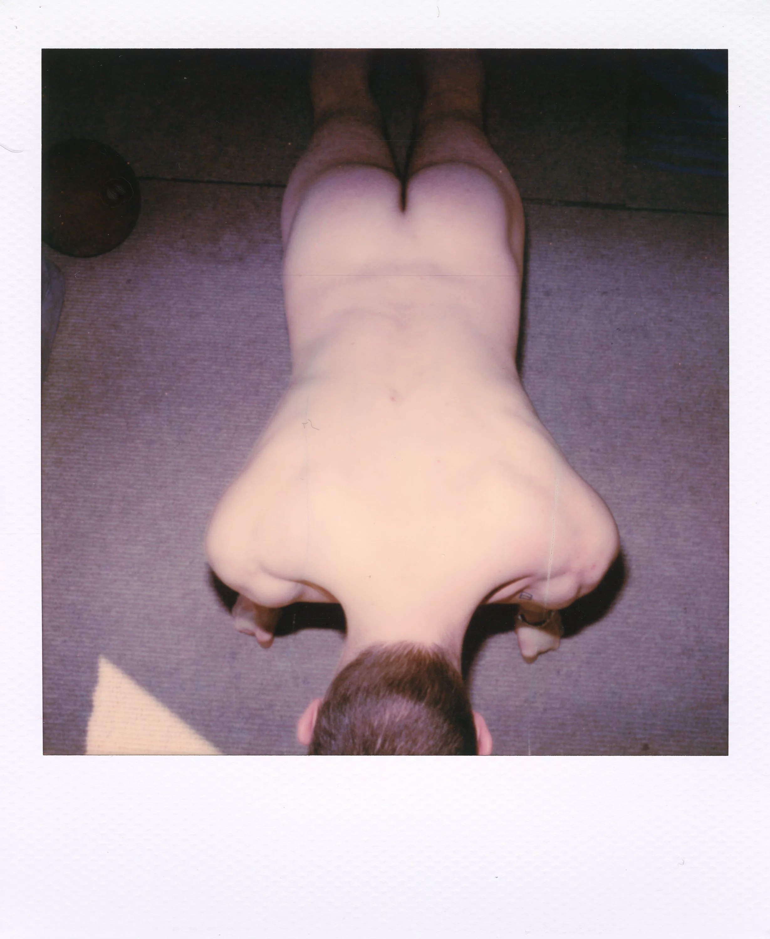 Top-down view of a person lying face down on the floor, with their back and buttocks exposed, hair short and brown, on a brown carpet with a beige mat nearby.