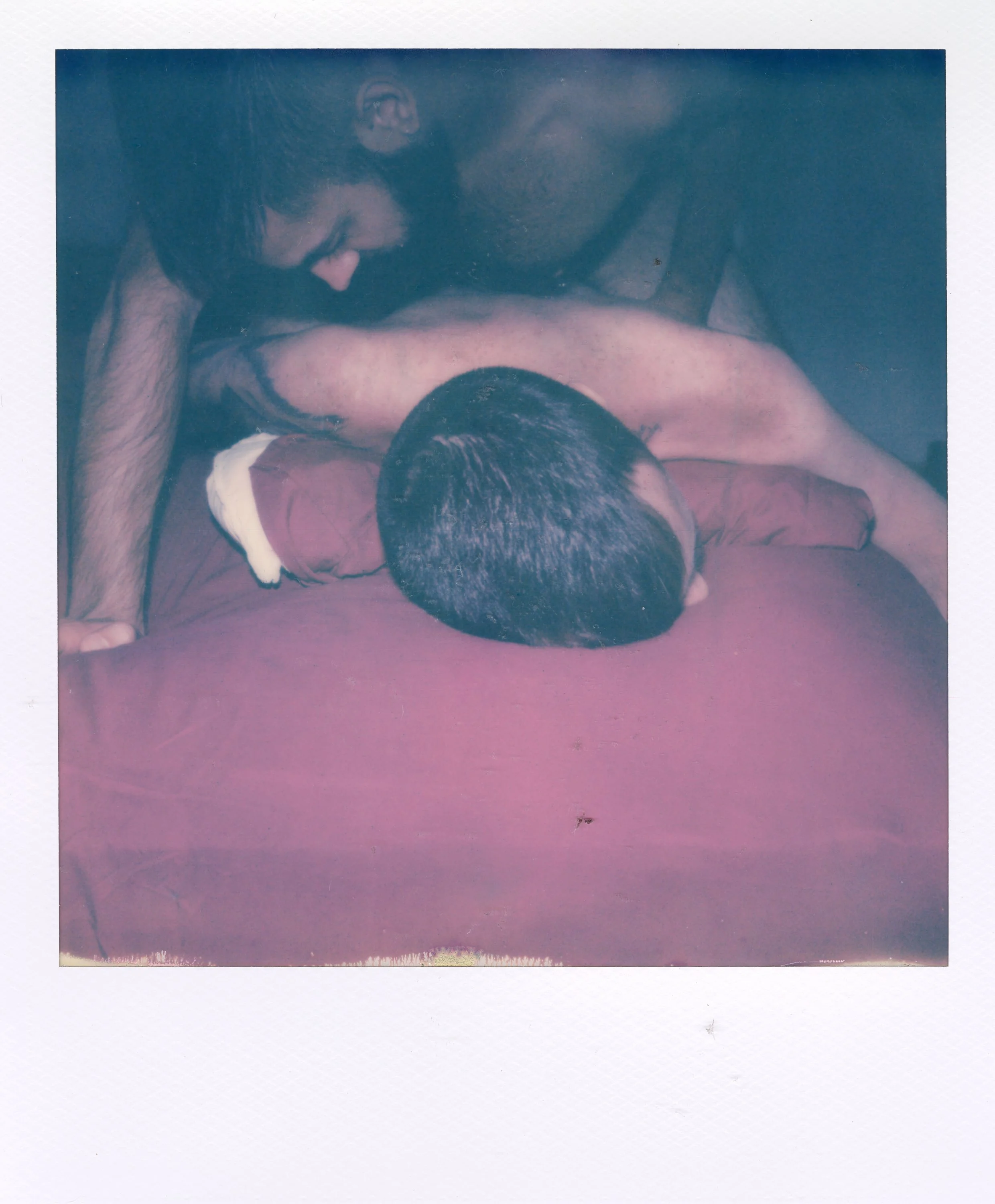 Two shirtless men are engaged in an intimate embrace on a bed with a burgundy sheet. One man is lying face down with his head turned to the side, while the other man leans over him. The scene appears to be a close, tender moment.