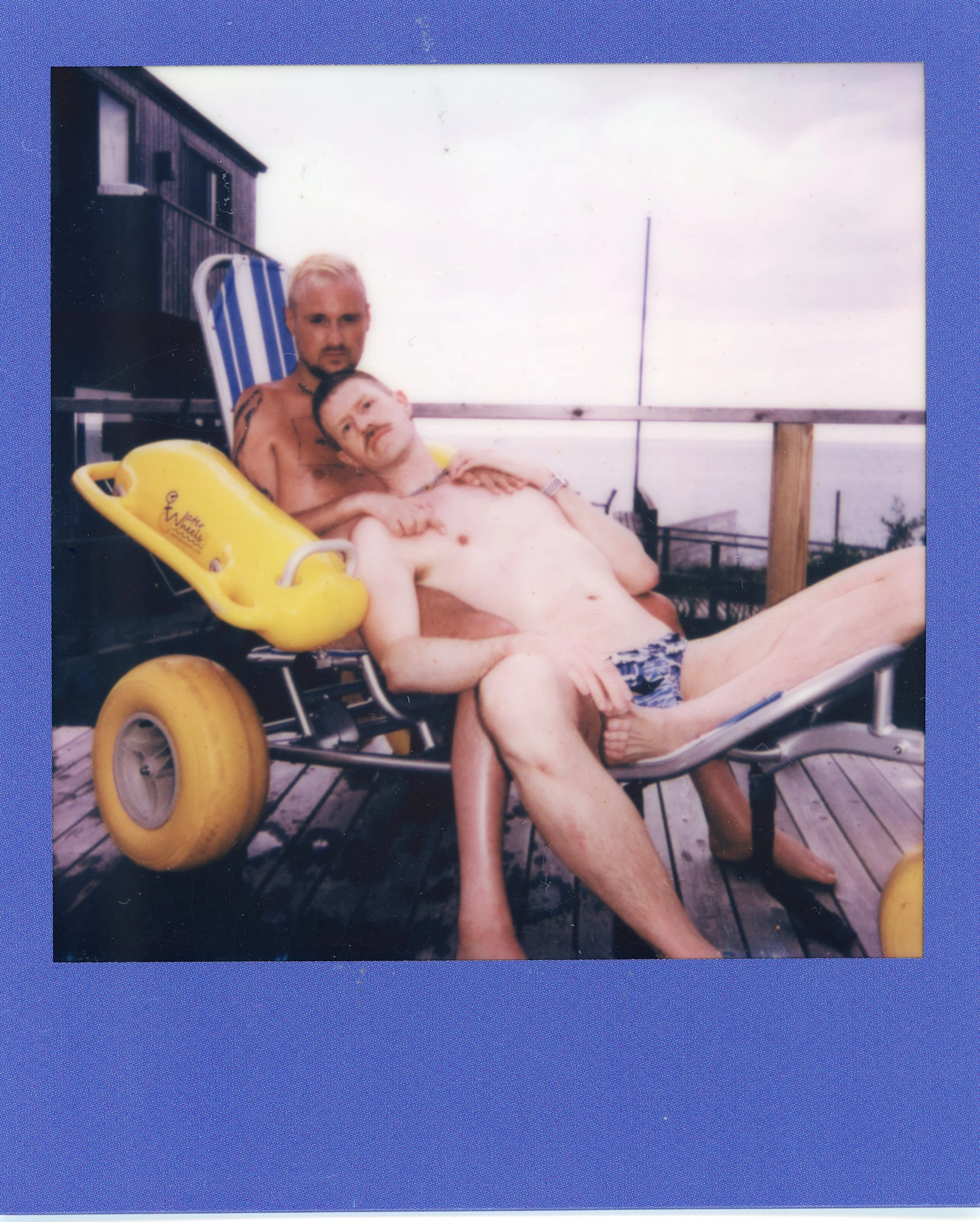 Two shirtless men relaxing on a wooden deck, one lying on a lounge chair and the other sitting beside him, with a cloudy sky in the background.