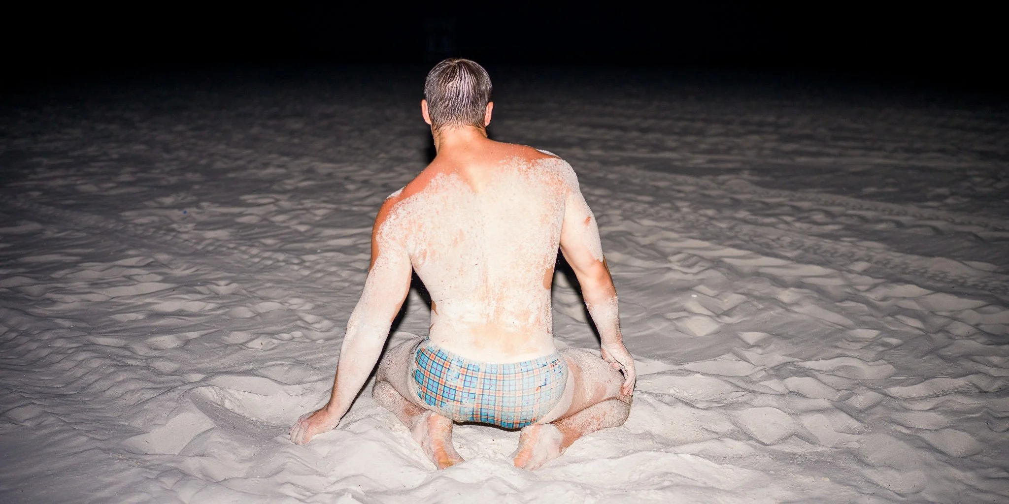 Man sitting on white sandy beach at night with sand covering his back and shoulders, wearing plaid shorts, facing away from the camera.