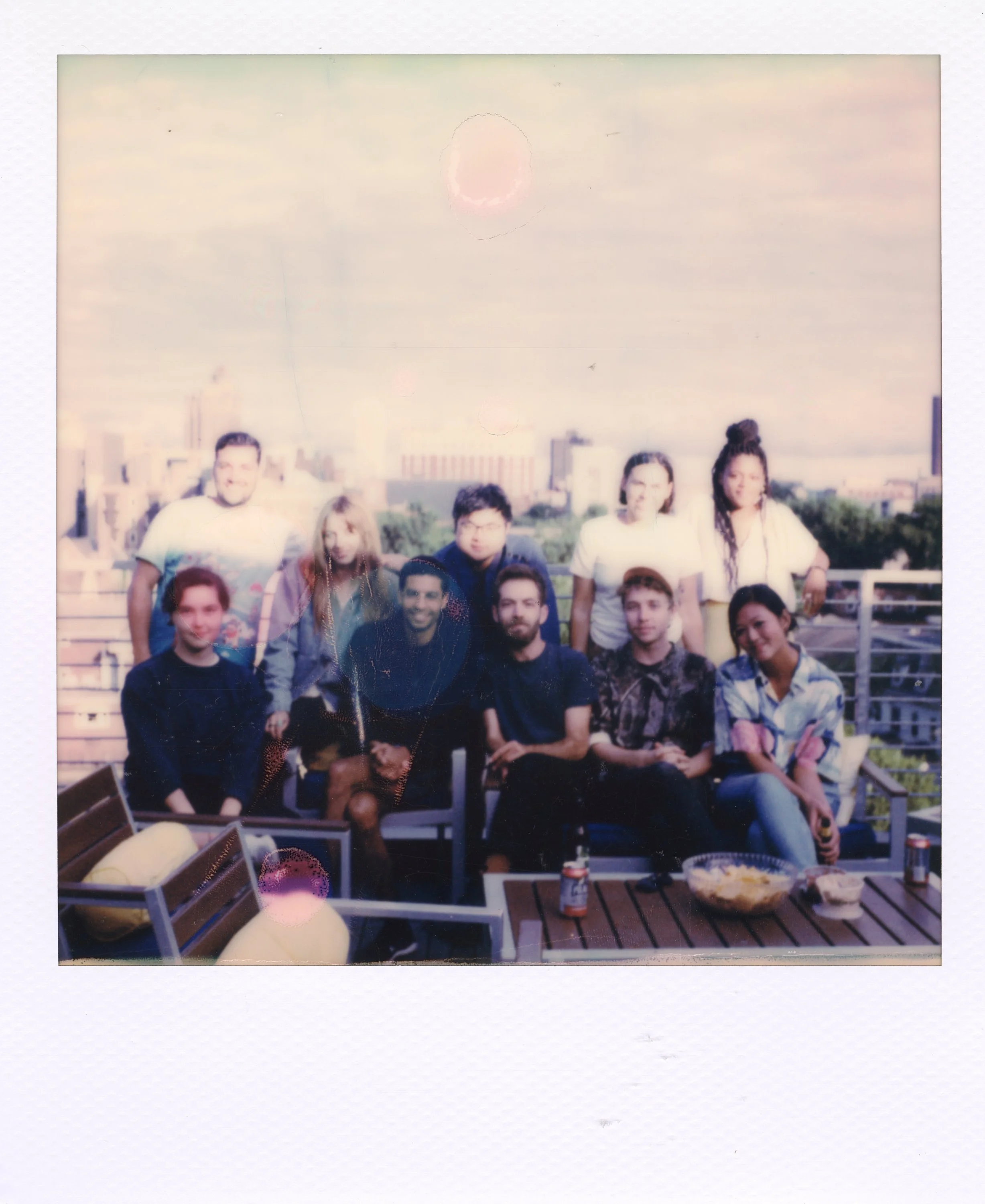 Group of ten friends on a rooftop with city skyline in the background, some sitting and some standing, with snacks and drinks on the table.