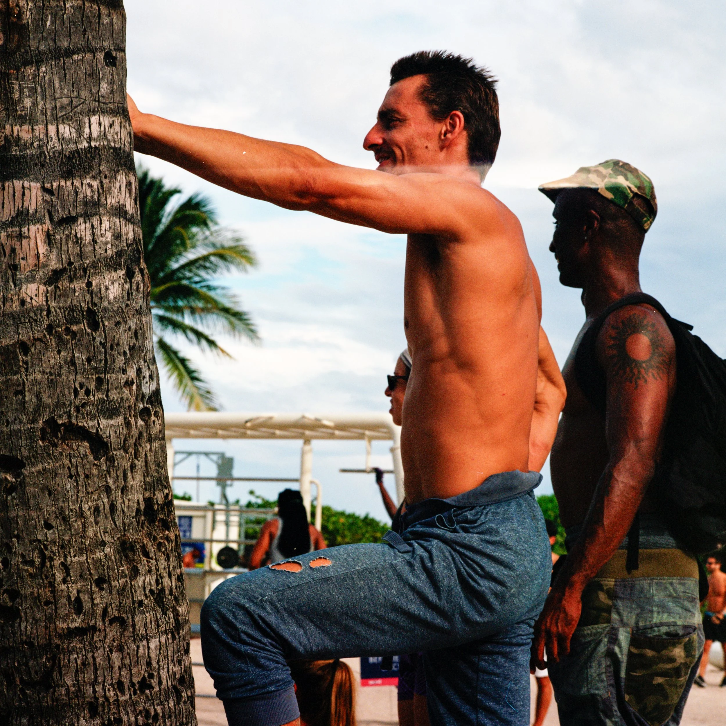 A shirtless man with dark hair doing a tree pose with one leg bent and foot resting on knee, against a background of palm trees and cloudy sky at an outdoor location, with other people in the background.