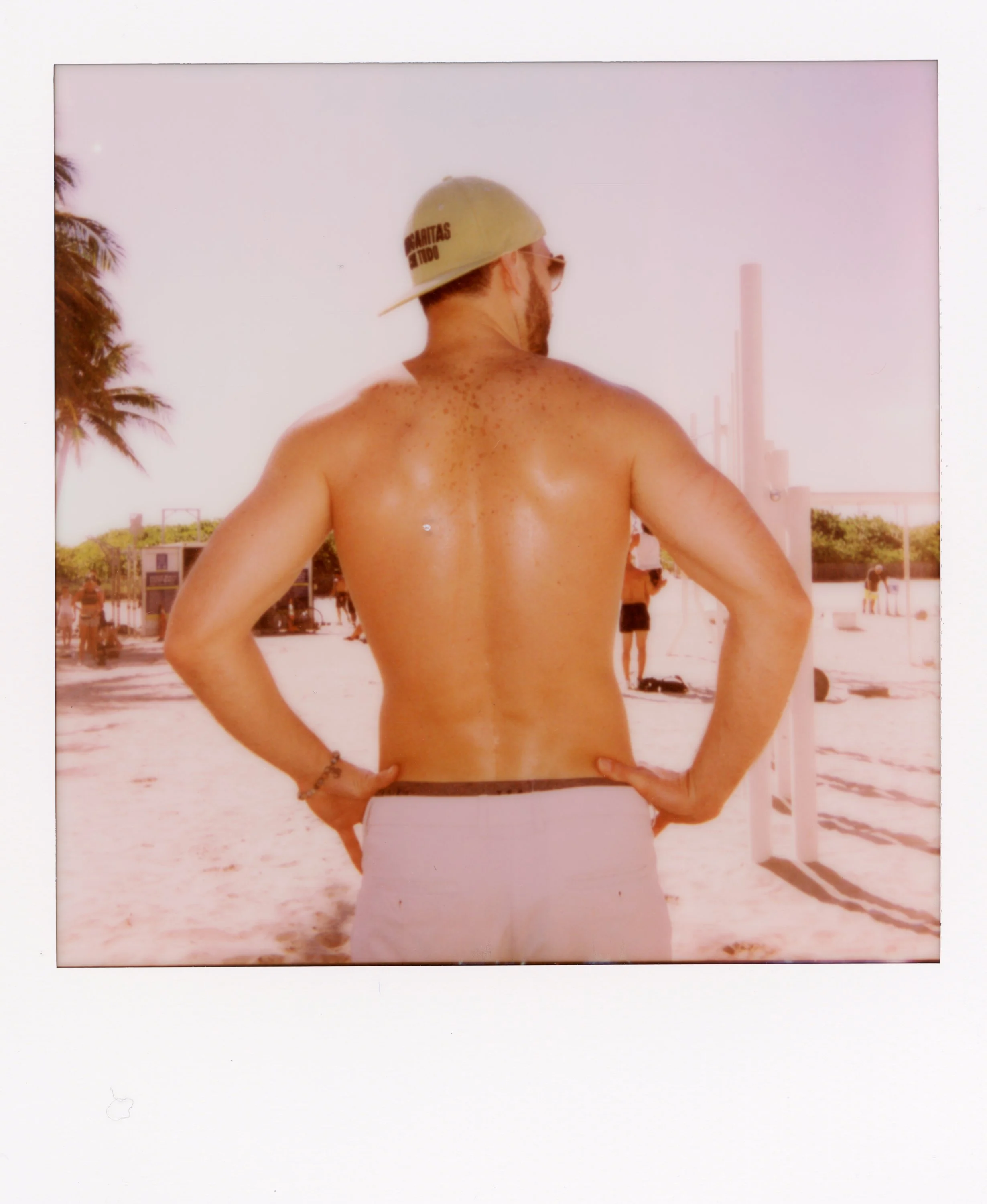 Back view of man shirtless on a beach, wearing a baseball cap and sunglasses, with palm trees and beach volleyball court in the background.