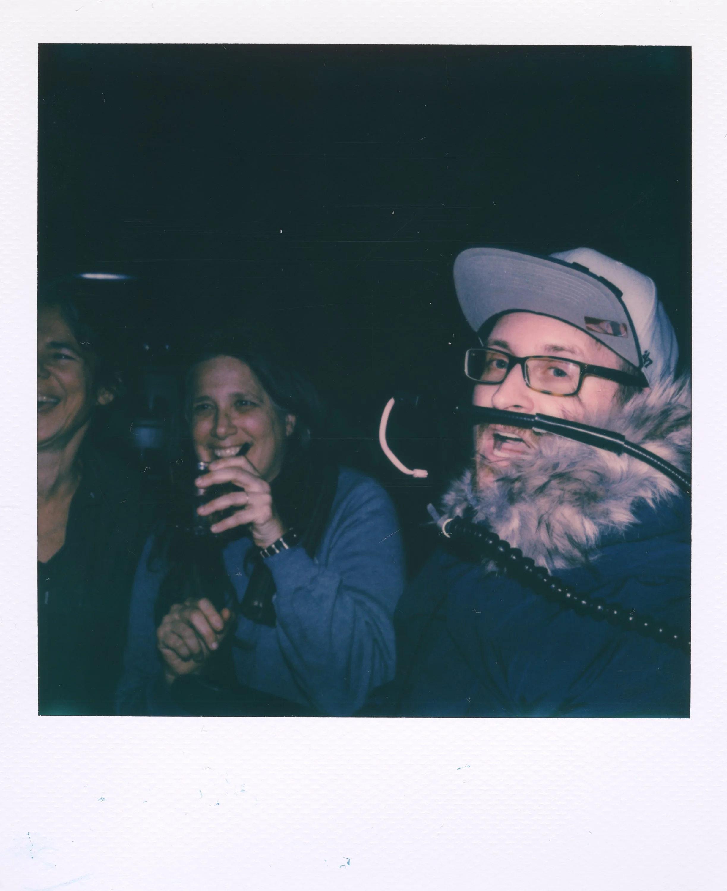 Group of people smiling and enjoying drinks at night, one person wearing a baseball cap and glasses, others visible in the background.