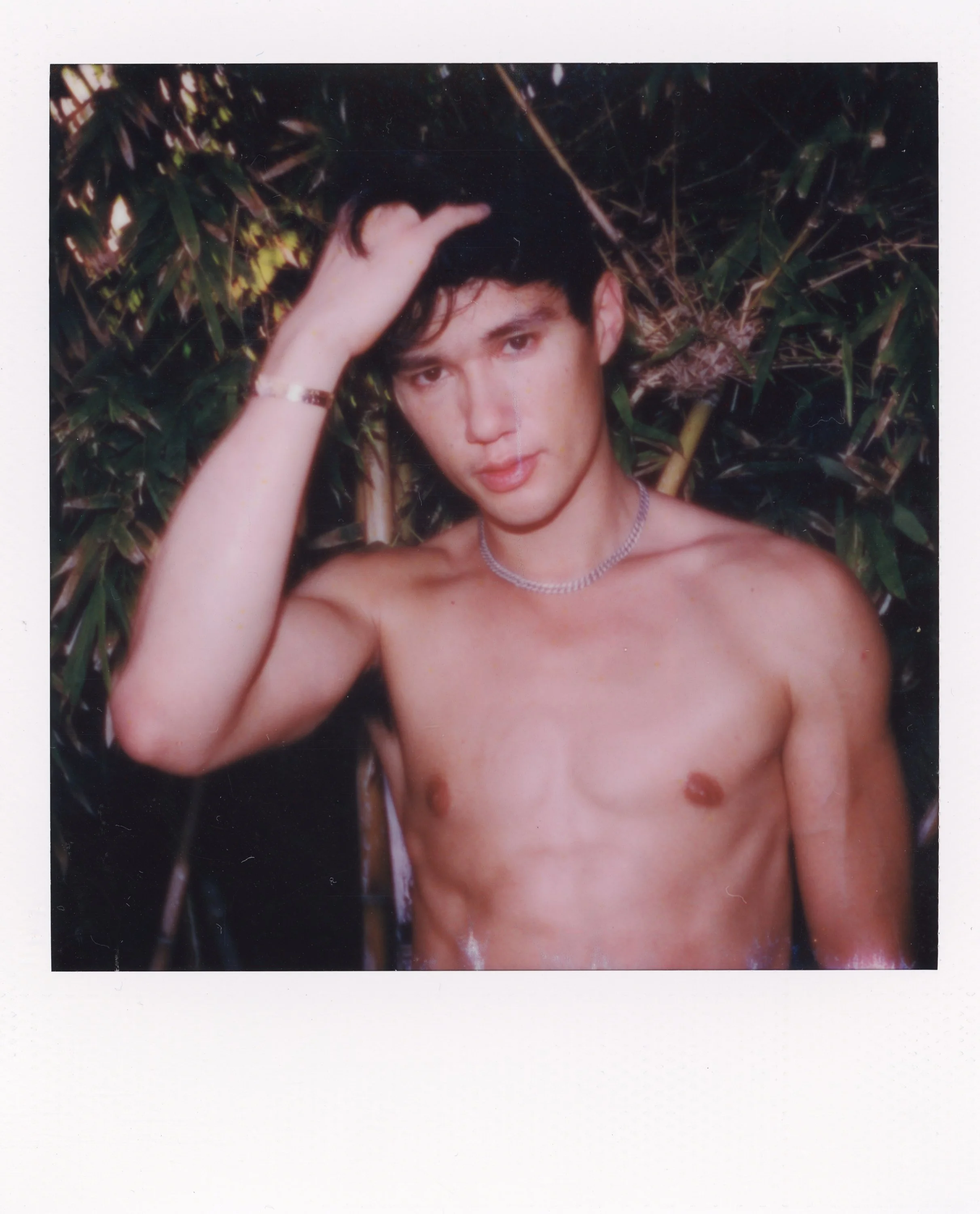A shirtless young man with dark hair standing in front of green foliage, wearing a necklace and a bracelet, looking at the camera with a relaxed expression and touching his hair.