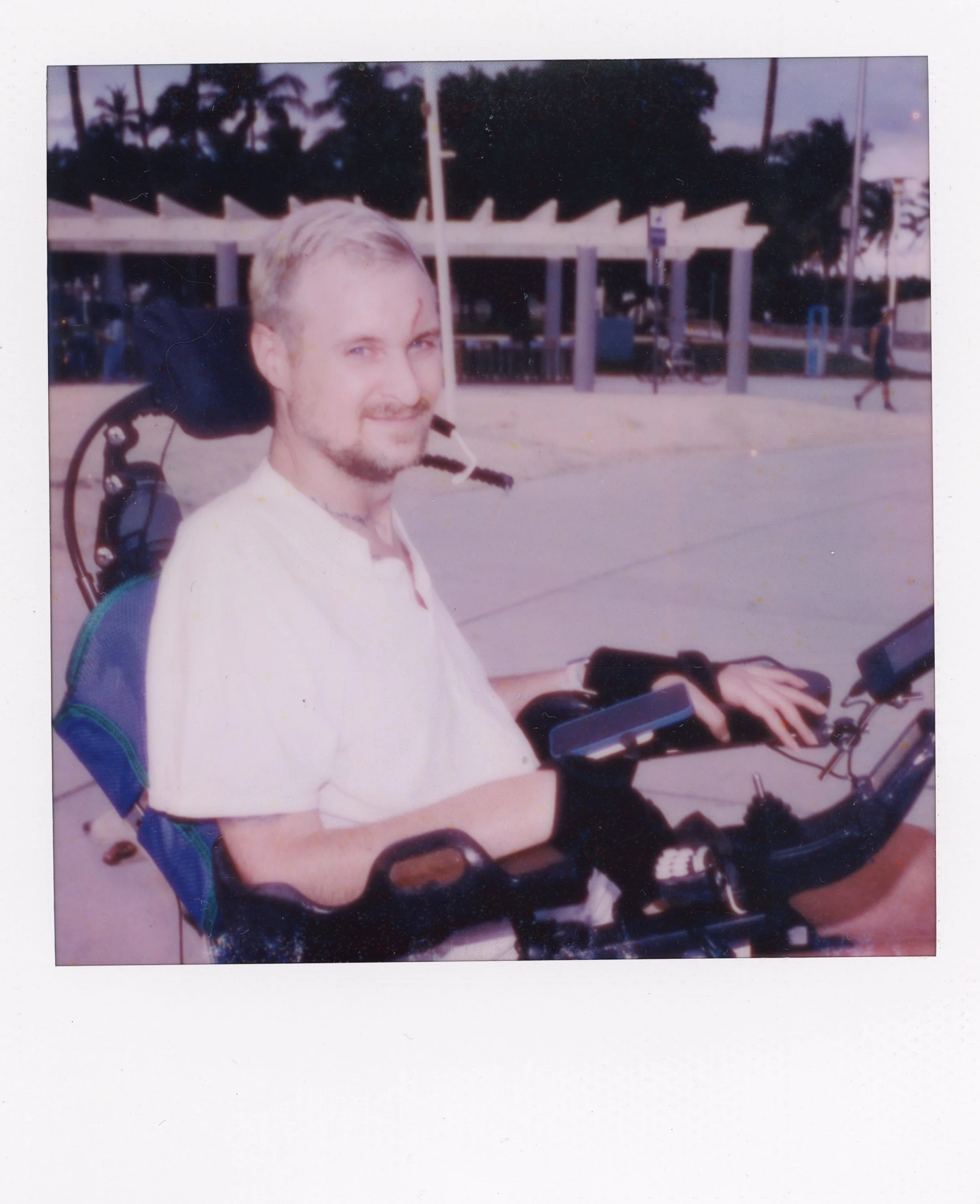 A man with short blond hair and a beard smiling while sitting on a motorized wheelchair outdoors, with trees and a pavilion in the background during dusk.