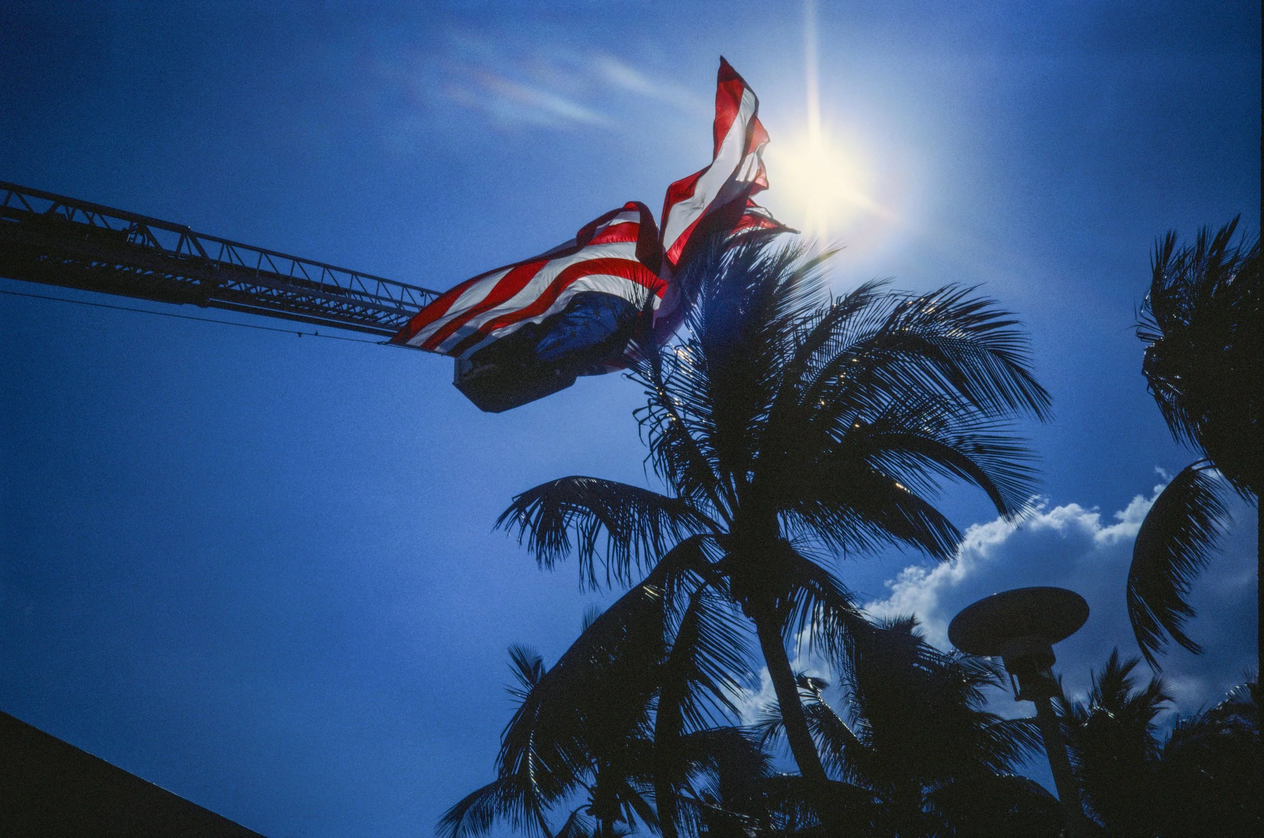 American flag flying on a sunny day with palm trees and a crane visible in the background.