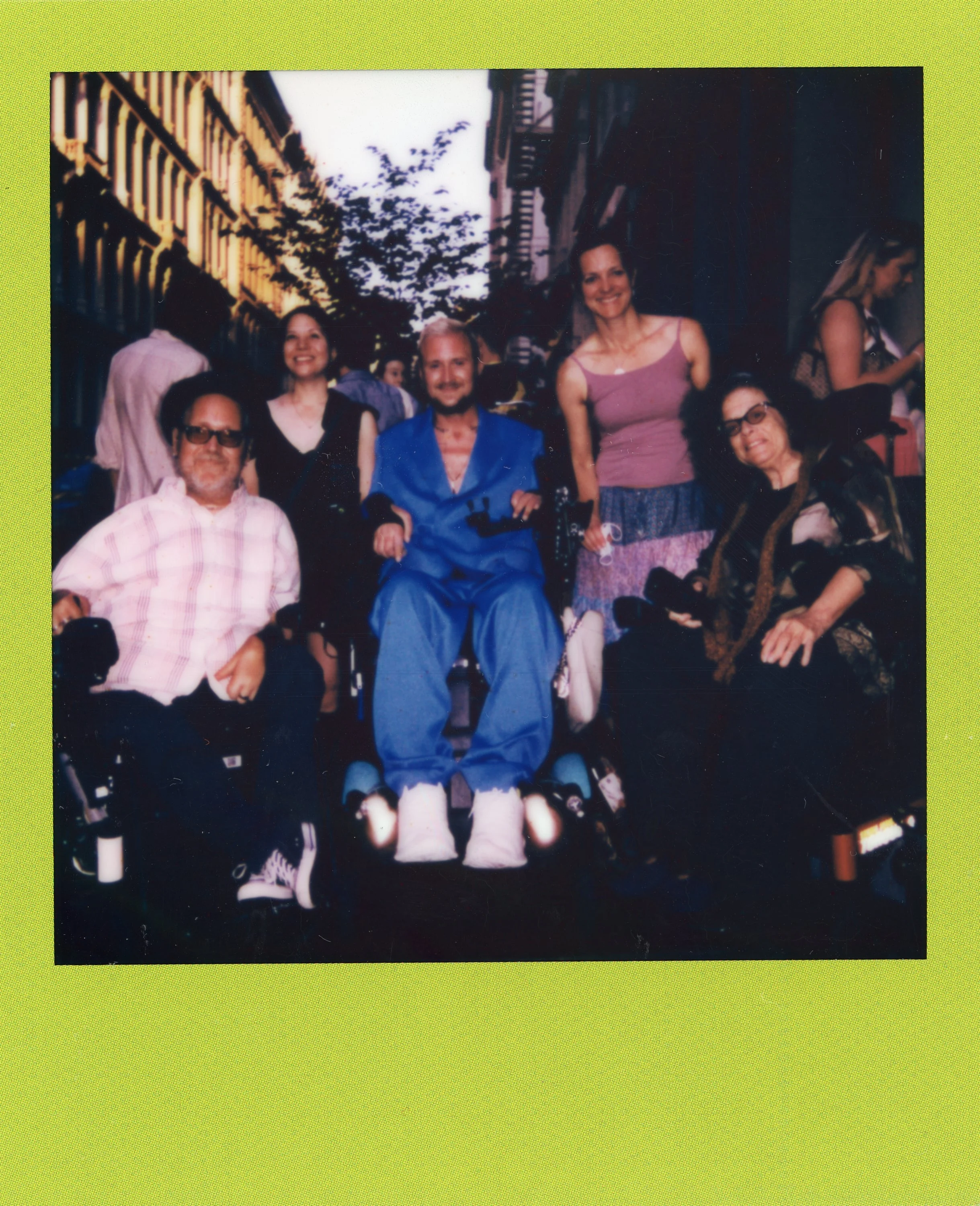 Group of six people, including two in wheelchairs, smiling outdoors on a city street with old buildings and trees in the background.