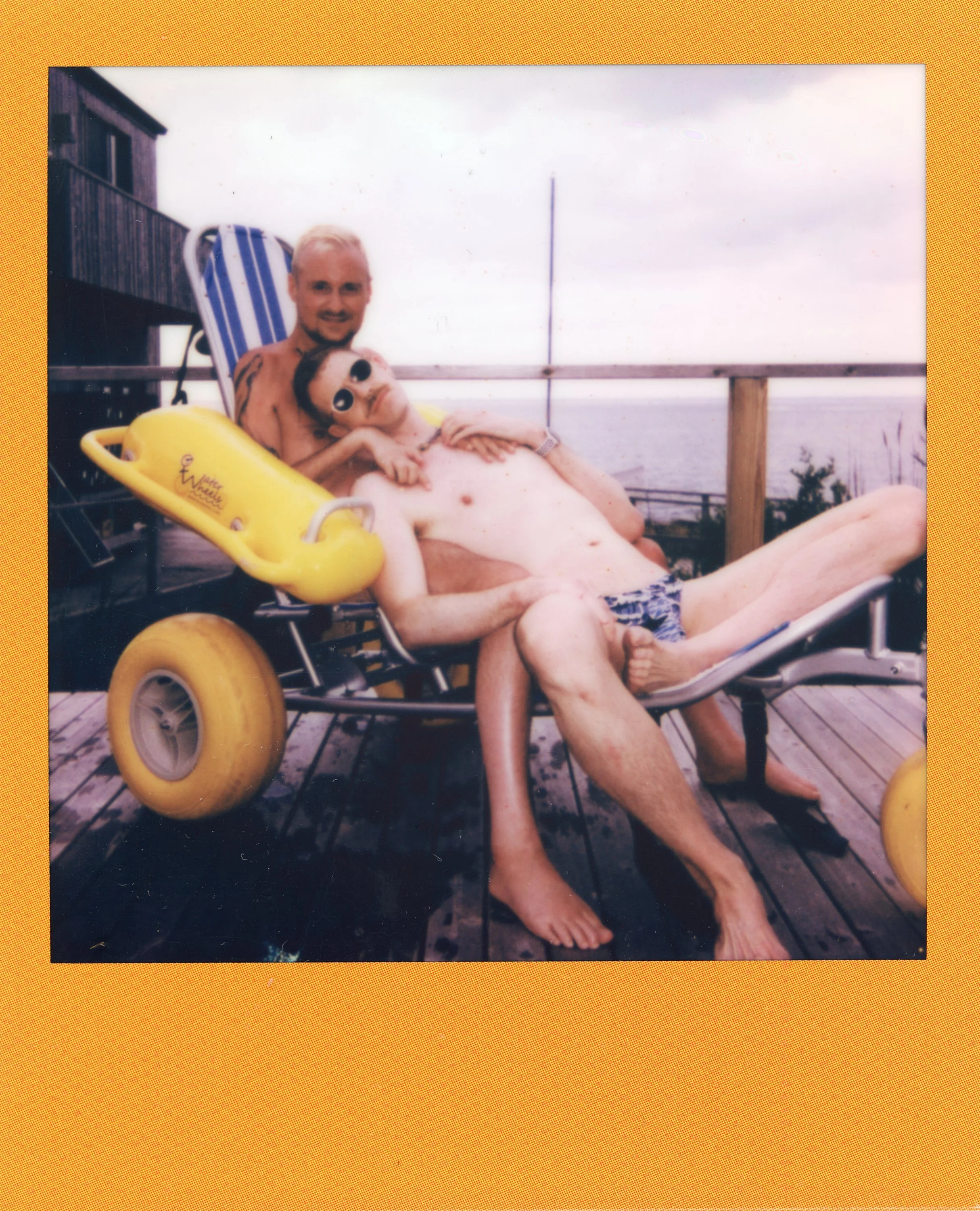 Two men sitting on a wooden deck with a view of the ocean in the background. One man is lying back on a lounge chair, wearing sunglasses, swim trunks, and is shirtless. The other man is sitting behind him, smiling at the camera. They seem relaxed and