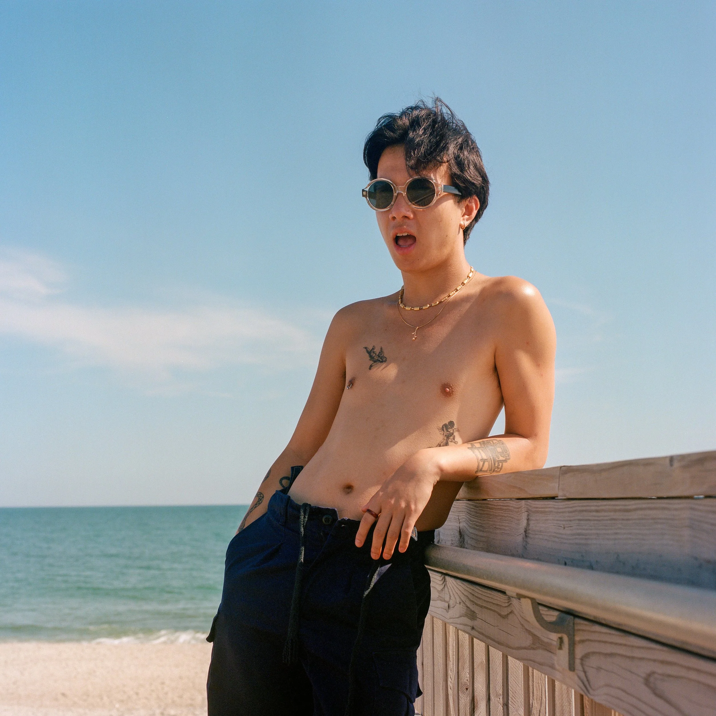 A shirtless person with sunglasses and tattoos leaning on a wooden railing at the beach.