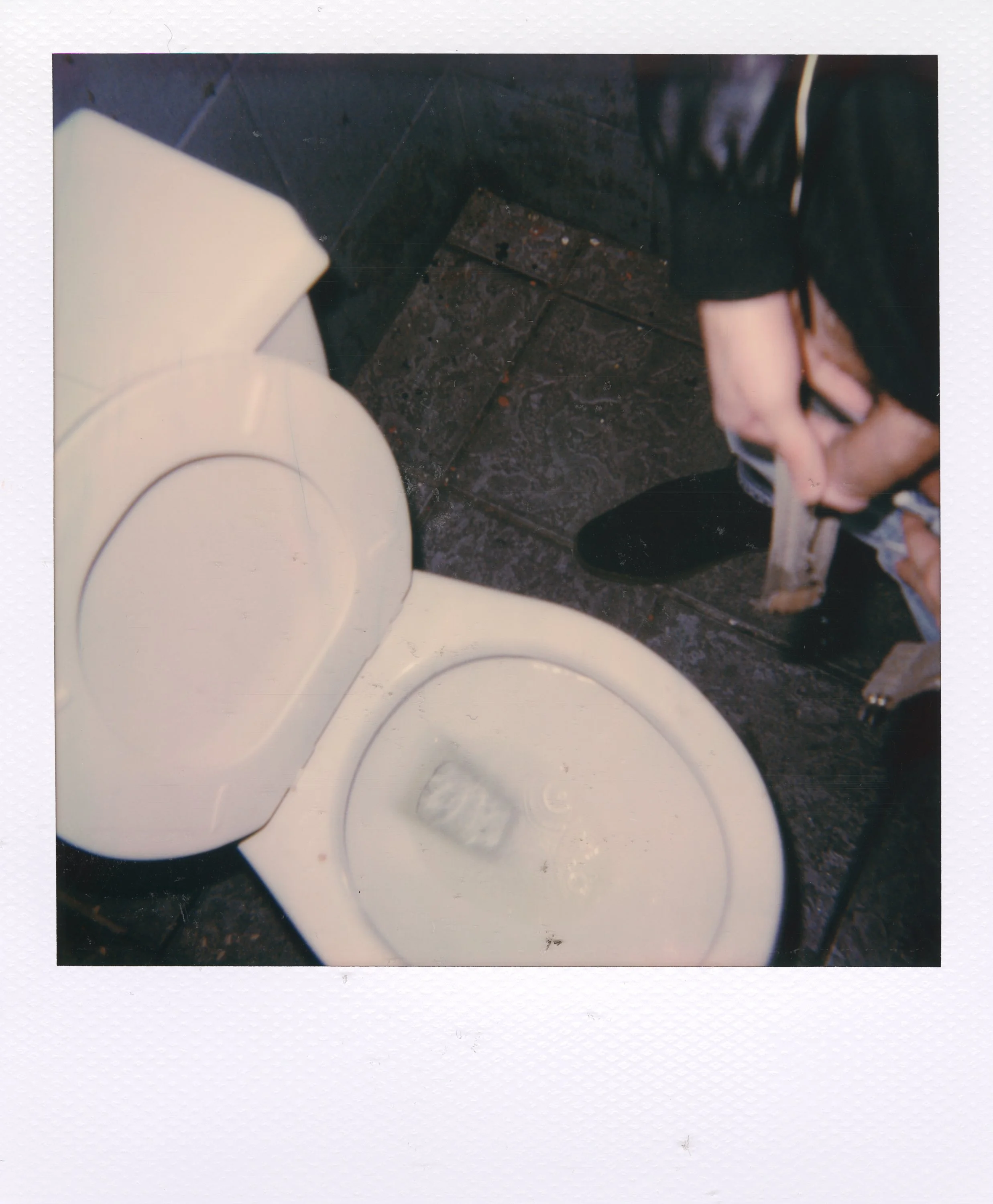 Public restroom with an open white toilet, a hand holding a spray bottle, and a person standing nearby wearing black shoes and clothing.