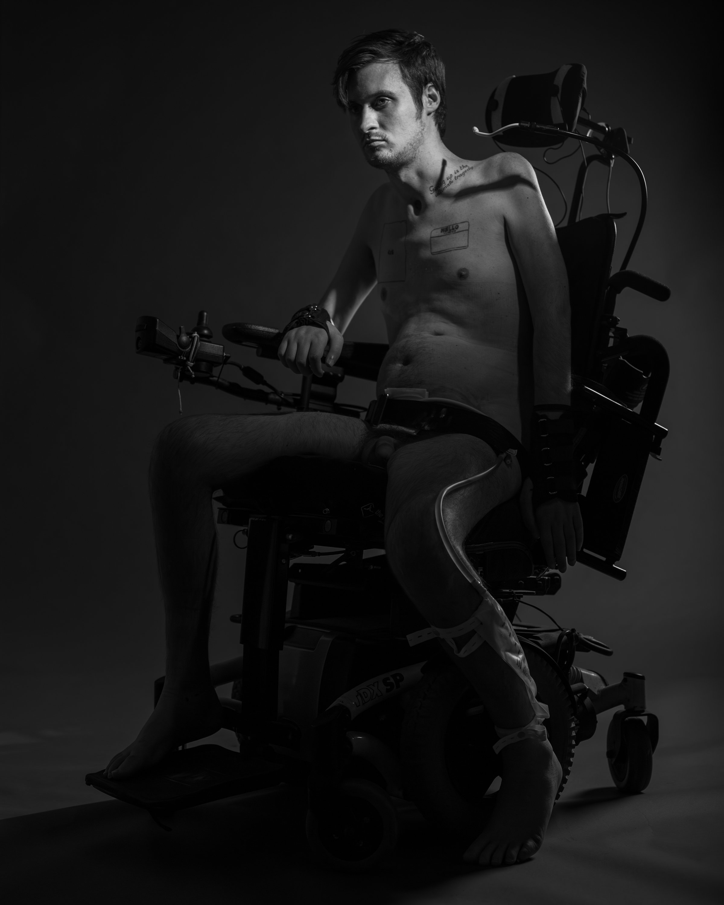 Black and white photo of a young man with a serious expression, sitting in a motorized wheelchair, shirtless, with tattoos visible on his chest and neck, holding the armrest with one hand, against a plain dark backdrop.