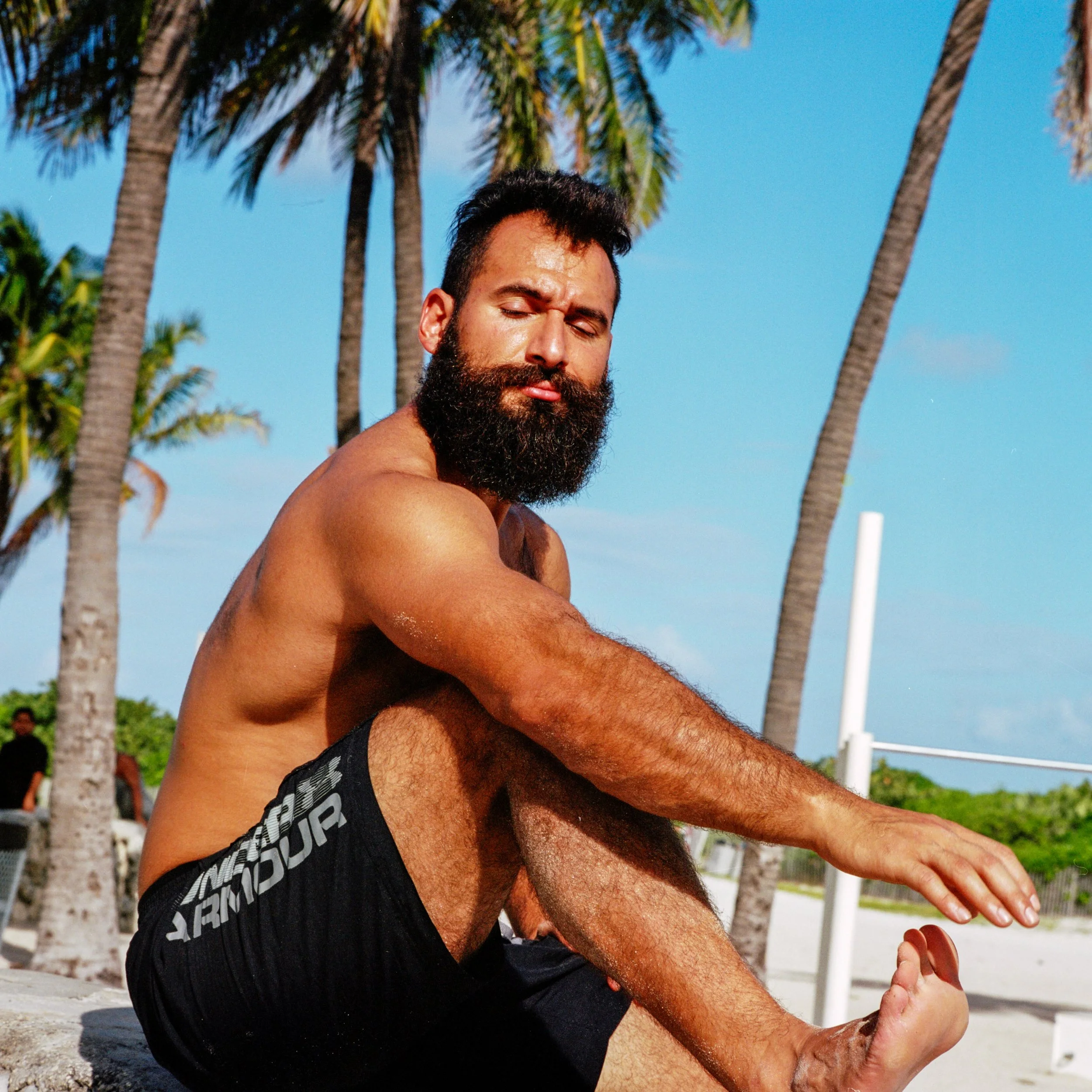 A man with a beard stretching on a beach with palm trees and a blue sky in the background.
