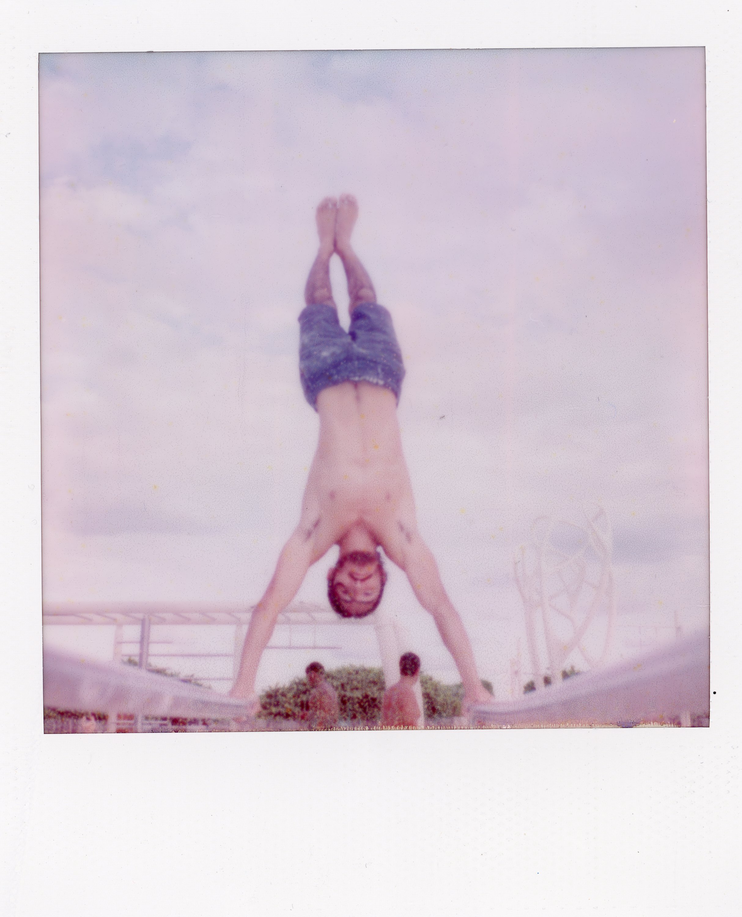 Person performing a handstand outdoors on a clear day, wearing shorts and glasses, with a Ferris wheel visible in the background.