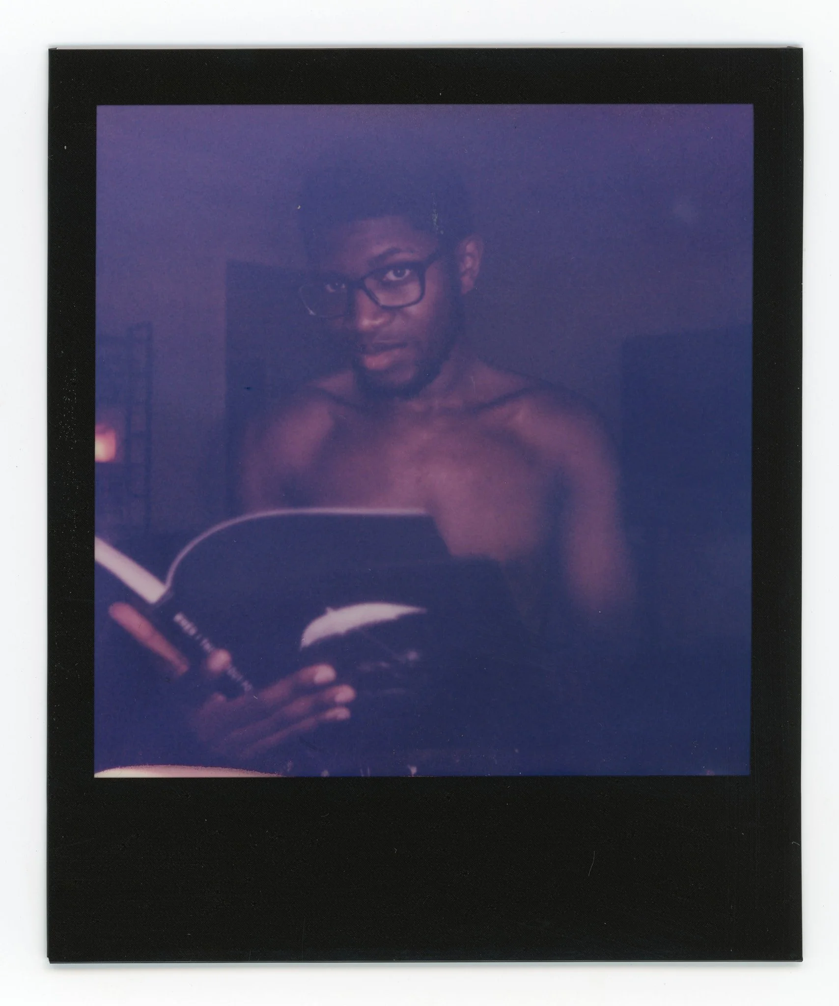 A shirtless young man with glasses holding a book and a pen, looking into a mirror in a dimly lit room.