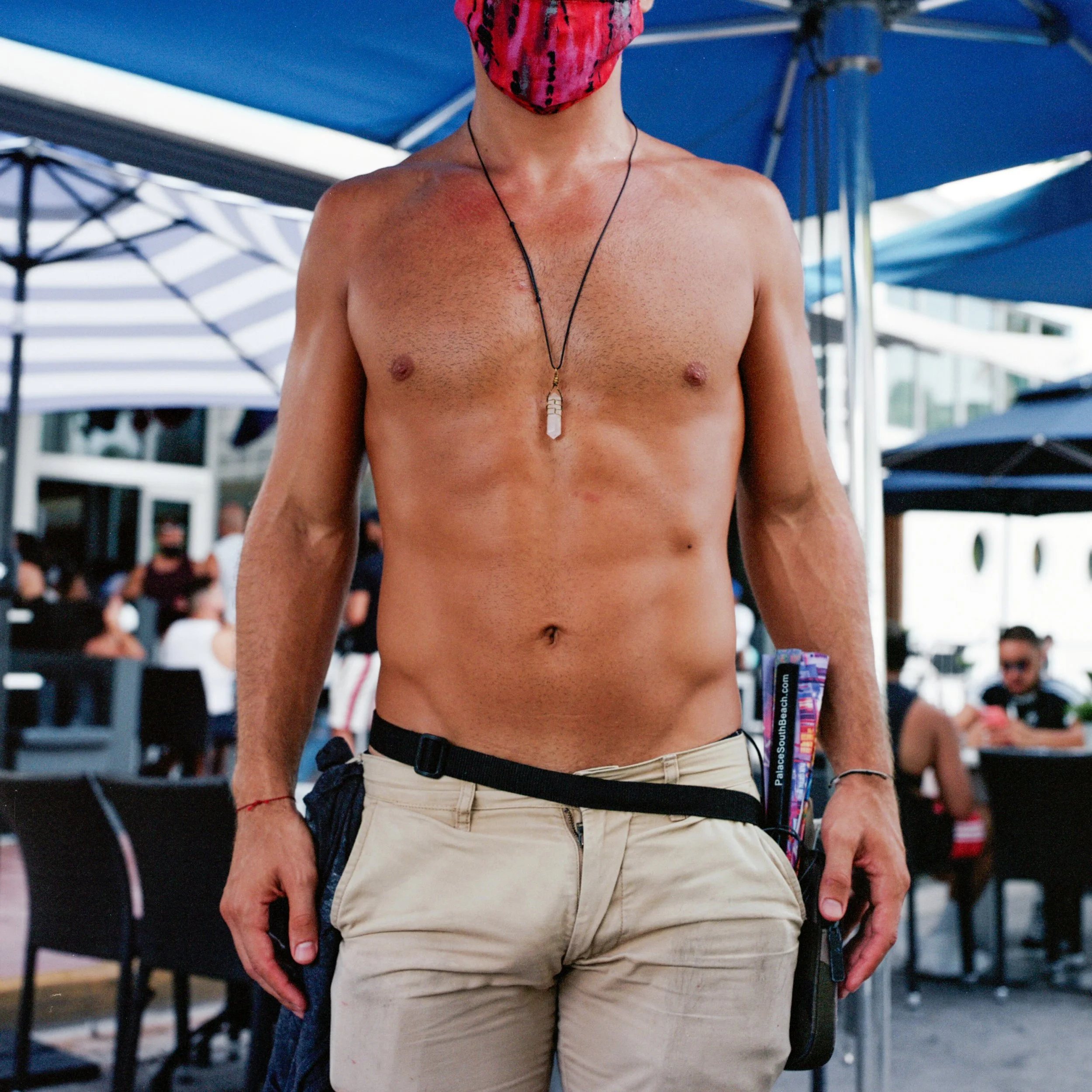 A shirtless man wearing beige pants, a pink face mask, and a necklace stands outdoors with a crowd in the background.