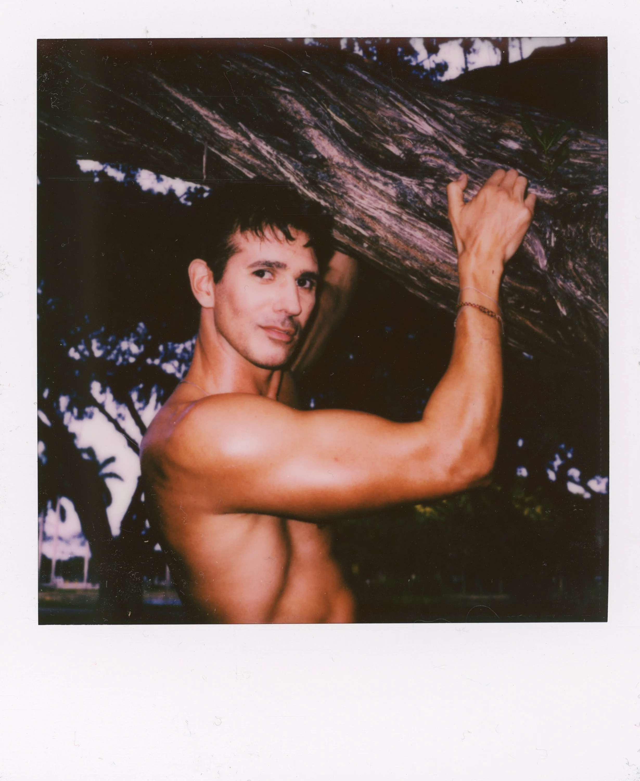 A shirtless man with dark hair and a slight smile, holding onto a large tree branch or trunk outdoors during dusk or dawn.
