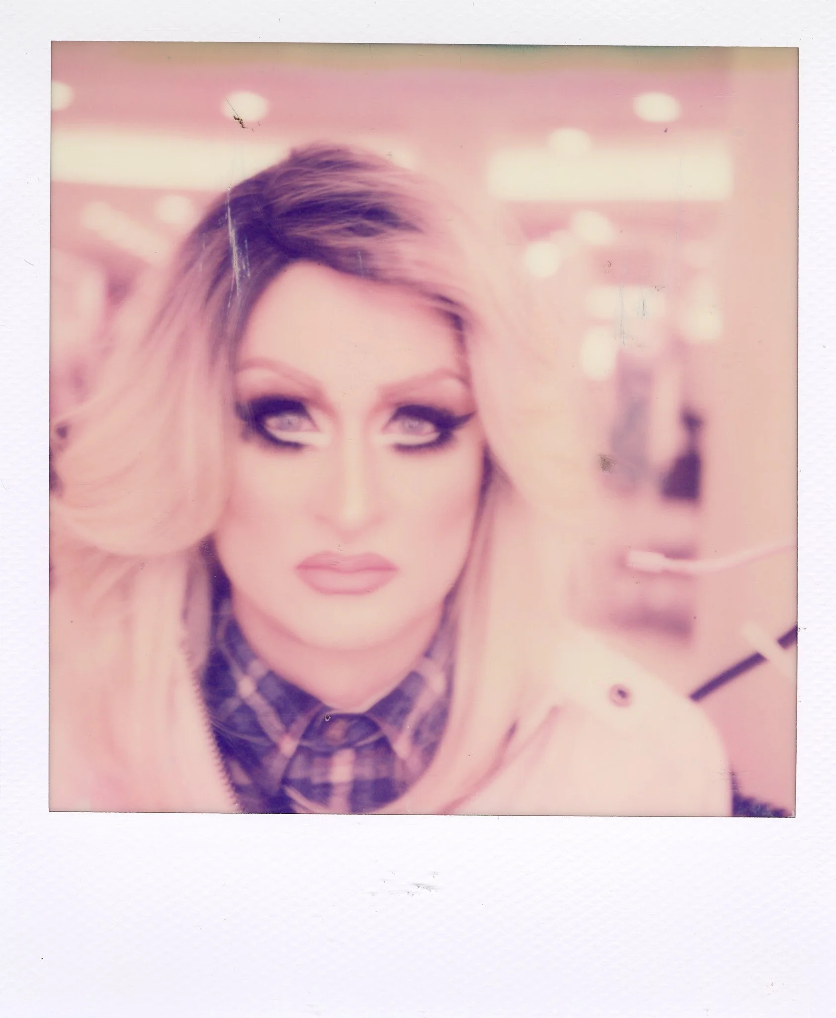 A woman with blonde hair, heavy makeup, and a plaid shirt, in a softly lit indoor setting, possibly a store or mall.