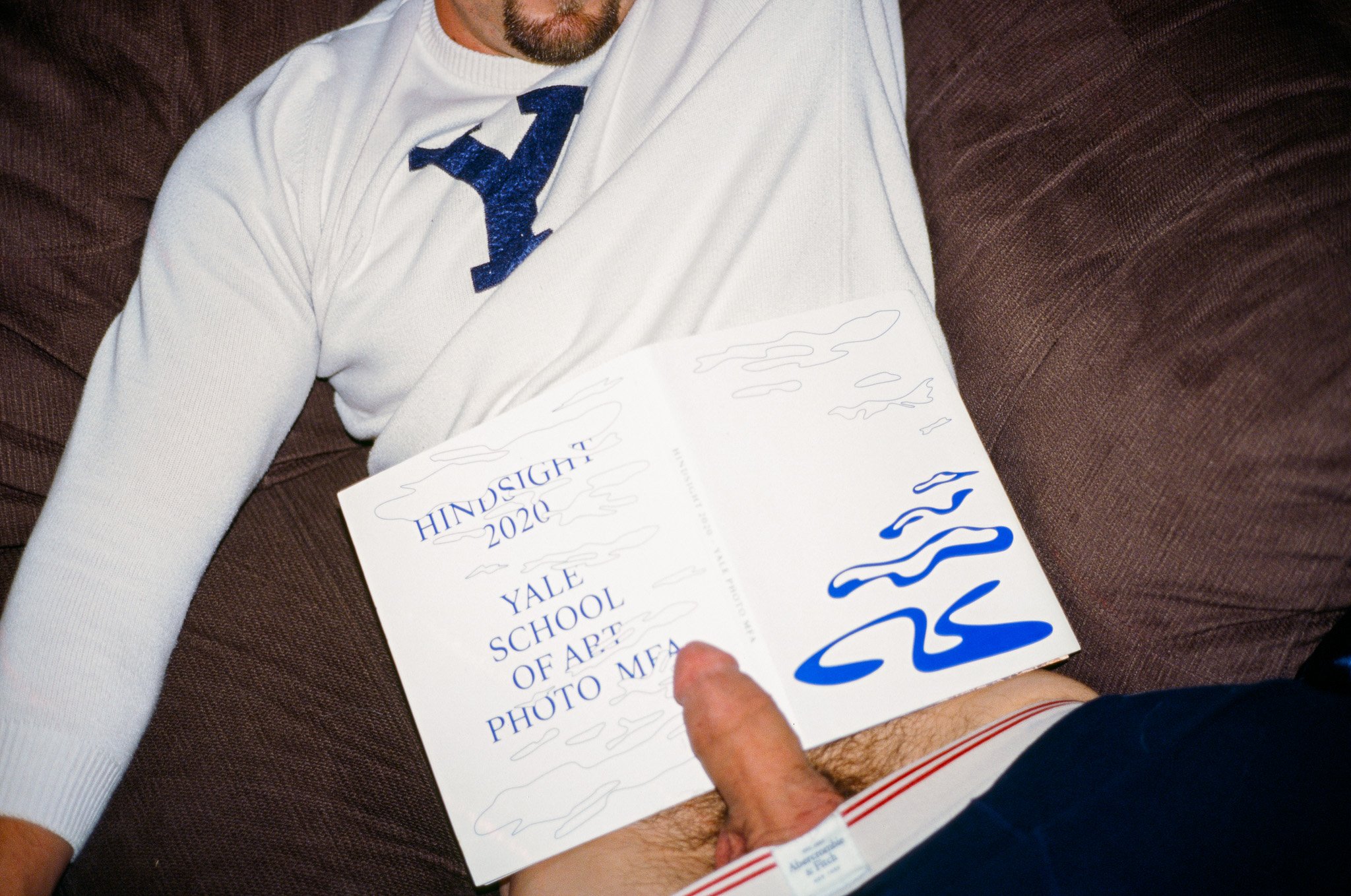 A person lying on a brown couch, wearing a white Yale sweatshirt with a blue letter Y. They are holding a booklet titled 'HINDSIGHT 2020 YALE SCHOOL OF ART PHOTO MFA' in their lap, and their other hand is resting on their thigh, which is dressed in s