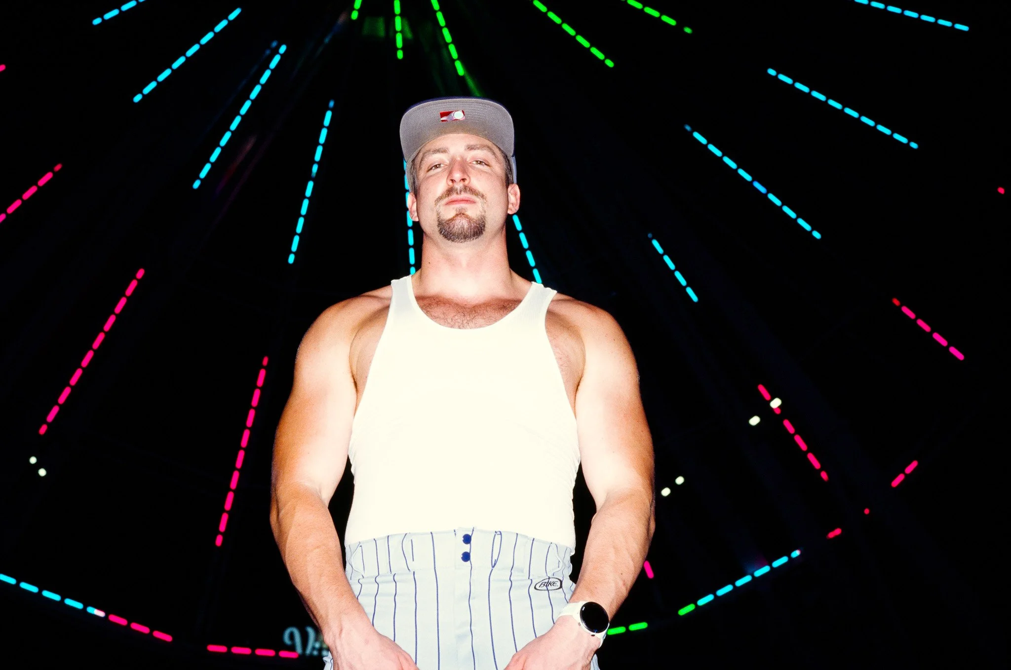 Man wearing a baseball cap, tank top, and striped shorts stands in front of a dark background with colorful, neon, dotted geometric lines radiating outward.