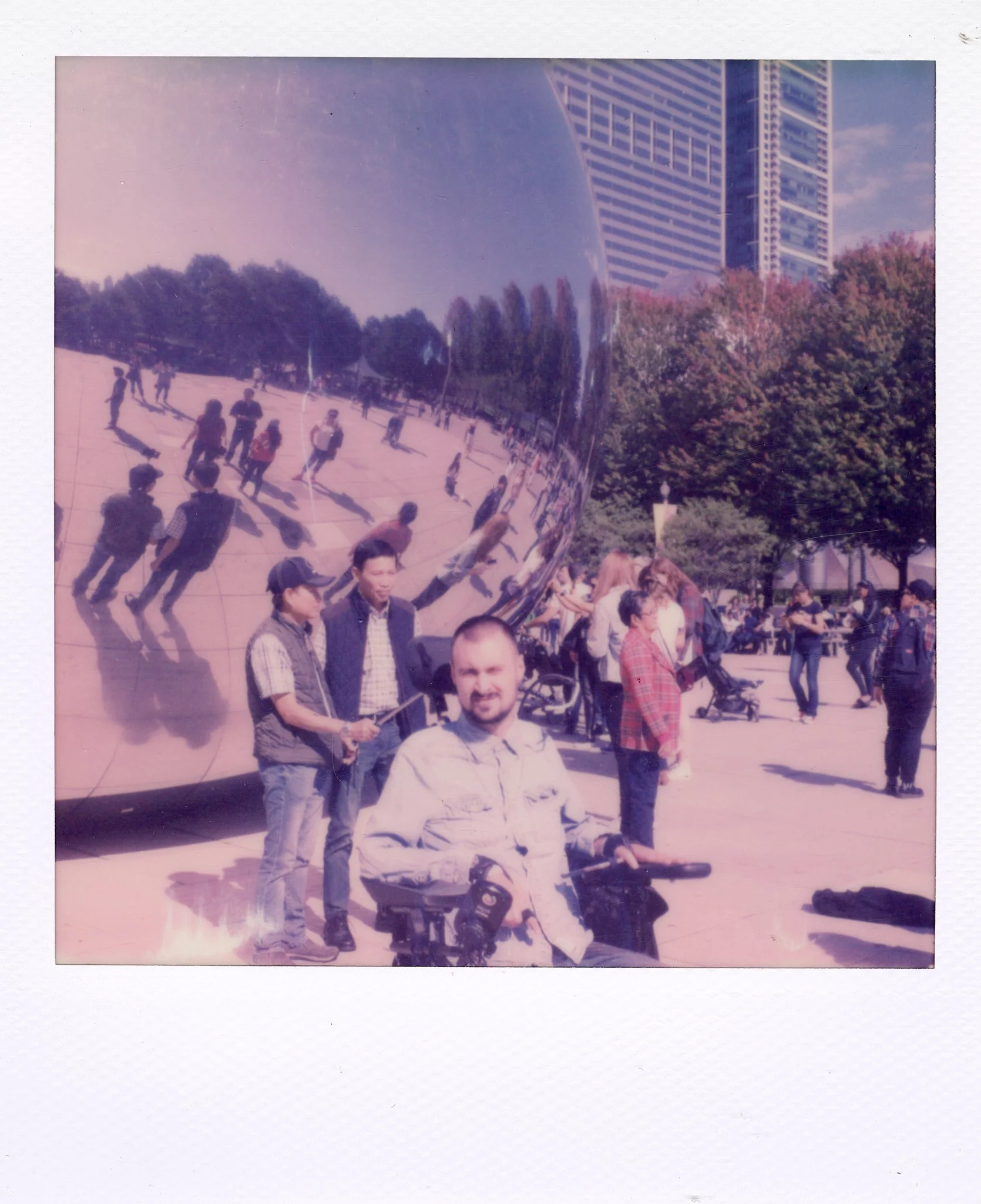 People gathered around the Cloud Gate sculpture in Millennium Park, Chicago, with buildings and trees in the background and the polished reflective surface of the sculpture showing distorted reflections of the scene.