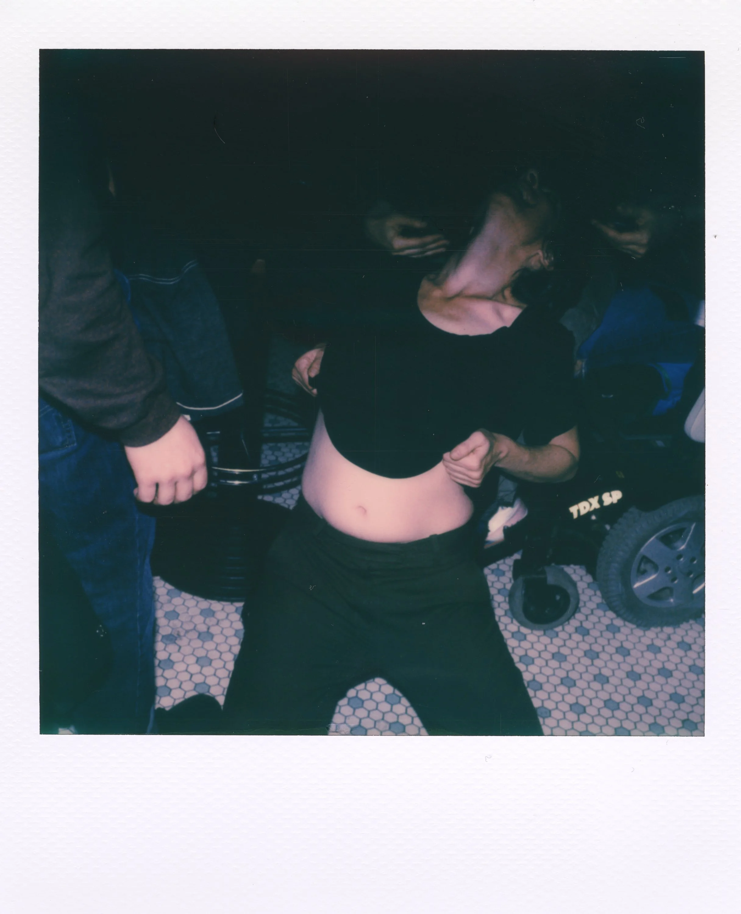 Person with black shirt lifting their shirt to reveal their abdomen, surrounded by other people, in a setting with tiled flooring and a wheelchair in the background.
