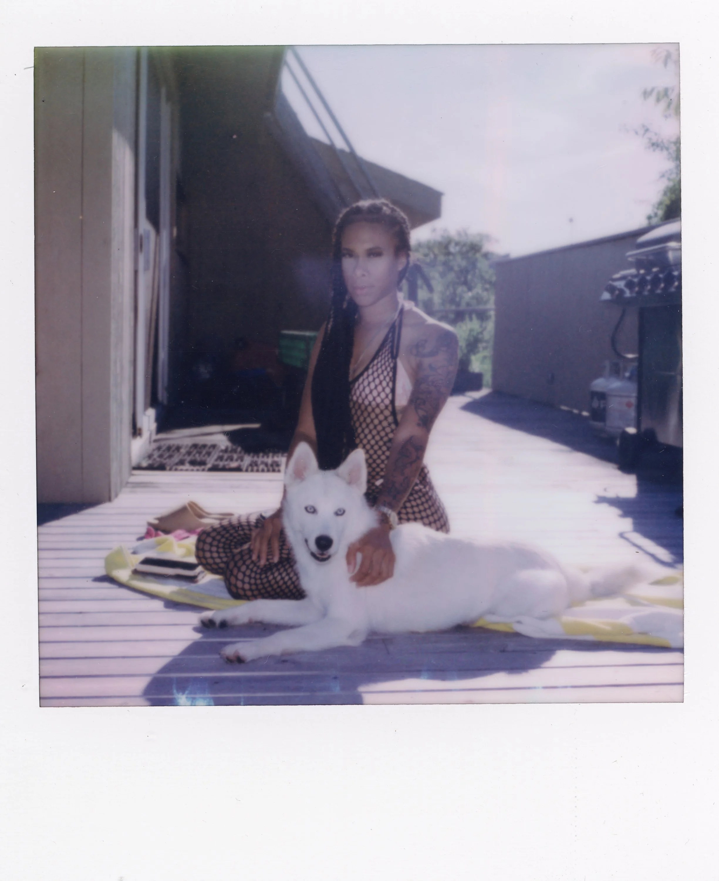 A woman with braided hair and tattoos sitting on a porch with a white husky dog, both looking at the camera.