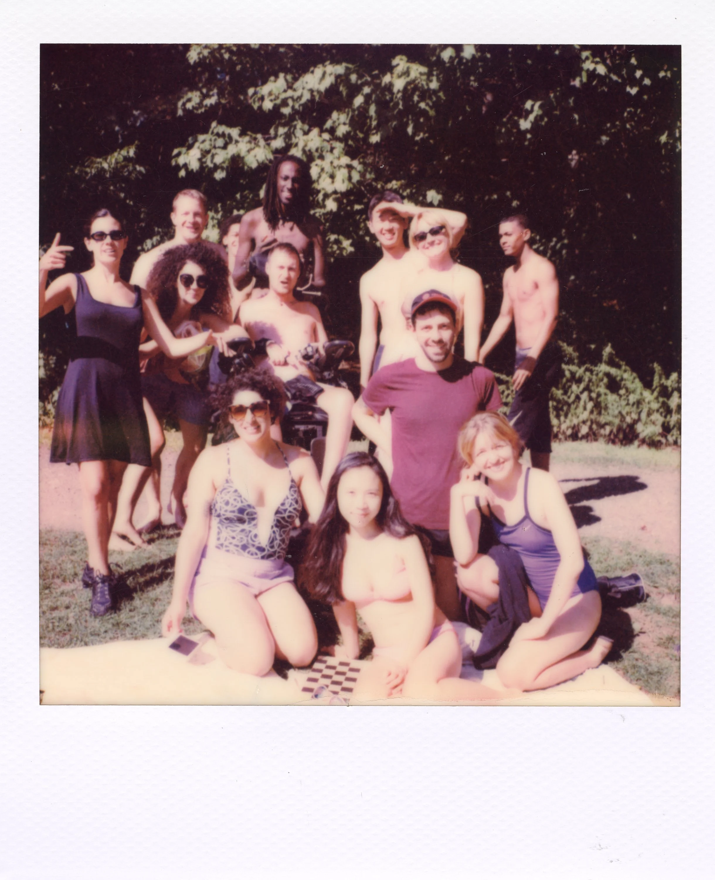 A group of people, some in swimsuits, gathered outdoors on a sunny day, posing for a photo with trees in the background.