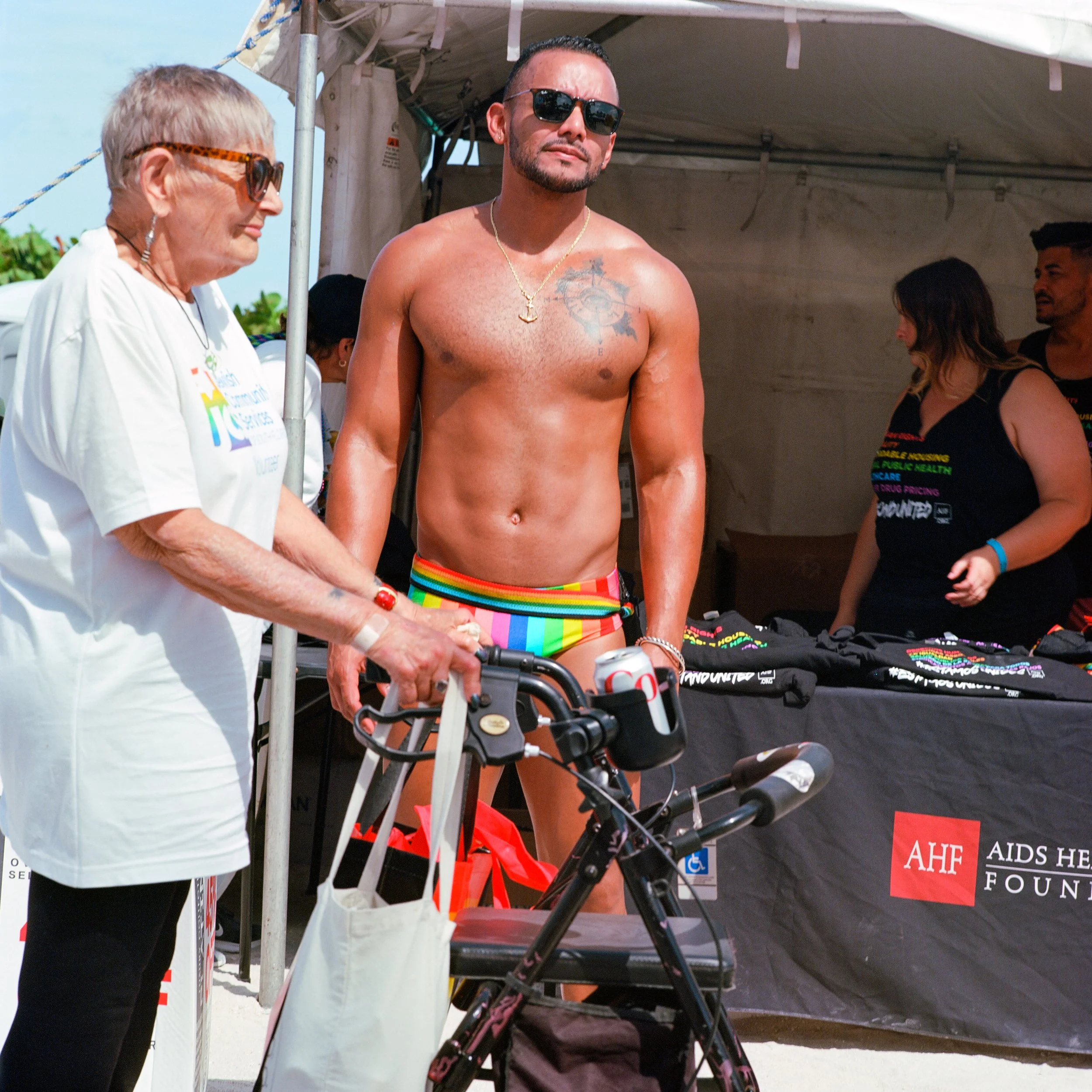 A shirtless man with rainbow-colored shorts and sunglasses standing next to an elderly woman with sunglasses, who is using a walker with a tote bag hanging from it. They are at a booth with a table covered in HIV/AIDS awareness merchandise, with a si