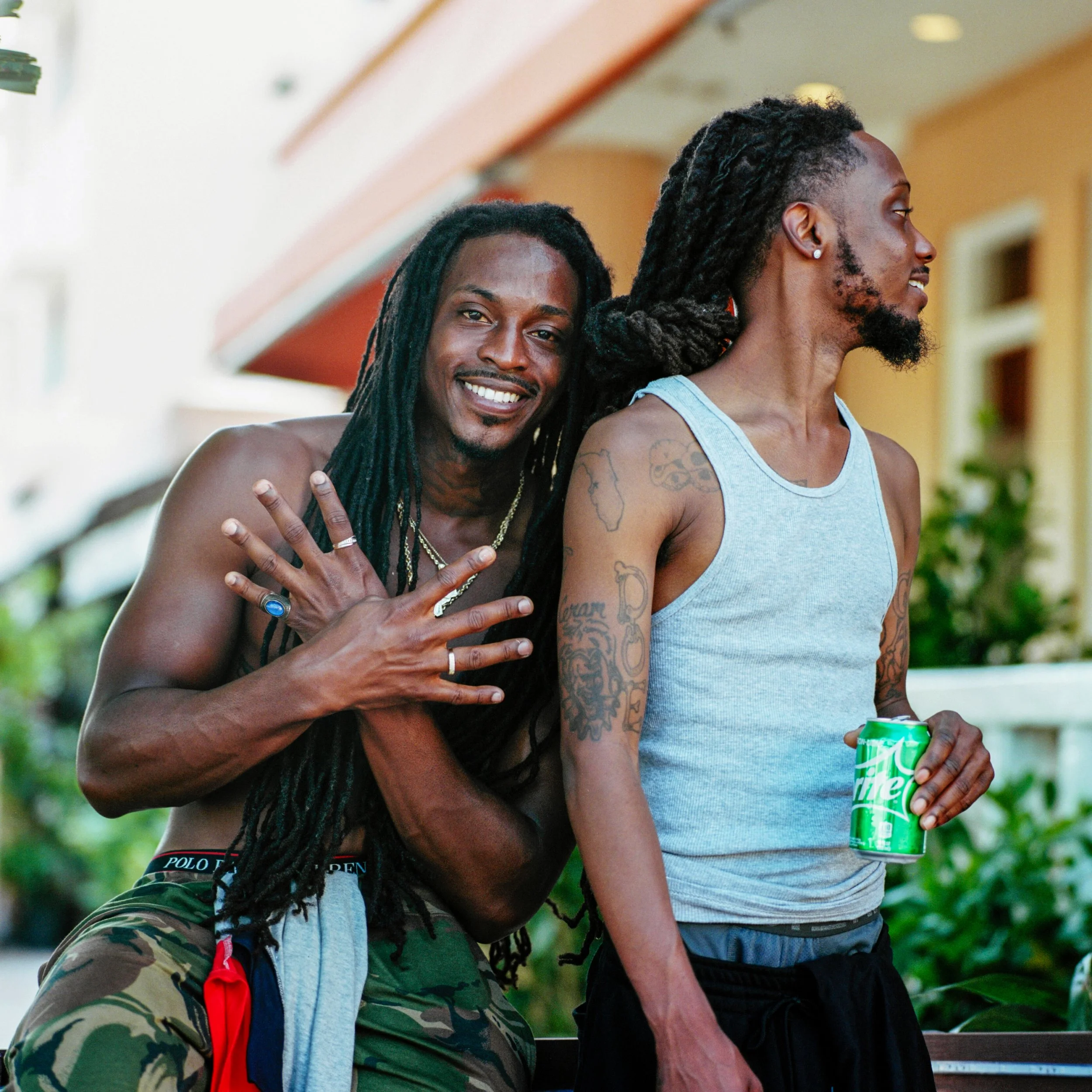 Two young men with dreadlocks outdoors, one smiling and showing his hand with rings, the other holding a Sprite soda can, both wearing casual tanks and shorts.