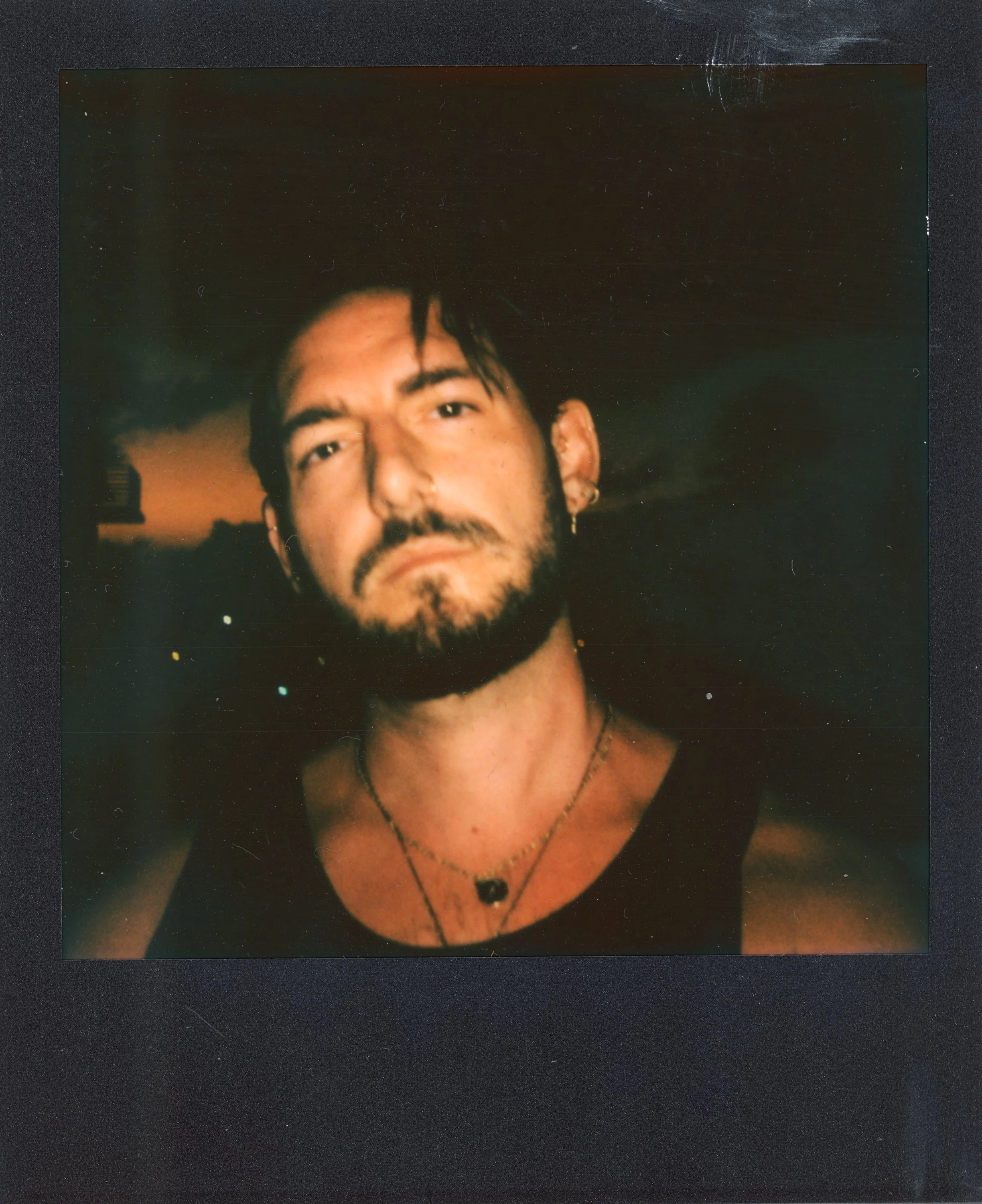 A young man with dark hair, a beard, and earrings, taking a selfie at night with a city skyline and a cloudy sky in the background.