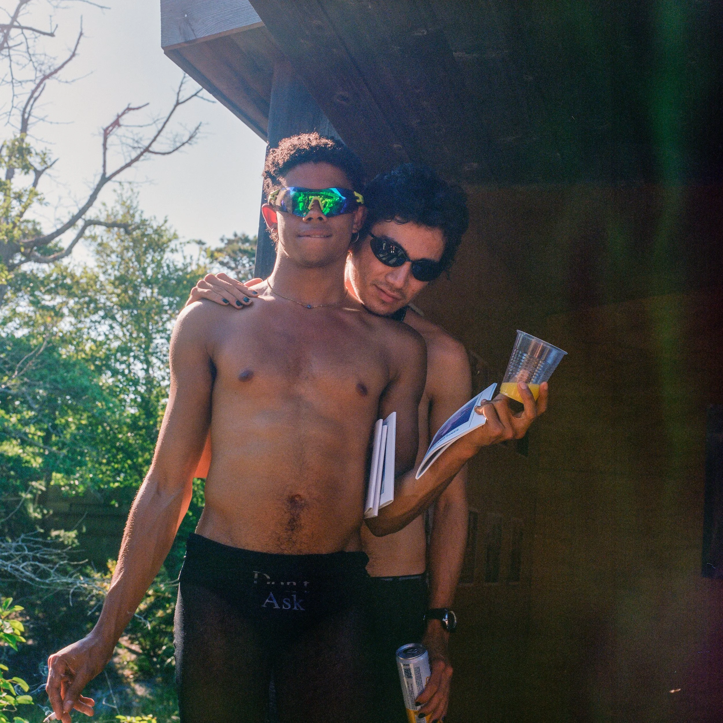 Two shirtless young men wearing sunglasses, standing outdoors, one holding a plastic cup and papers, with trees and a wooden structure in the background.
