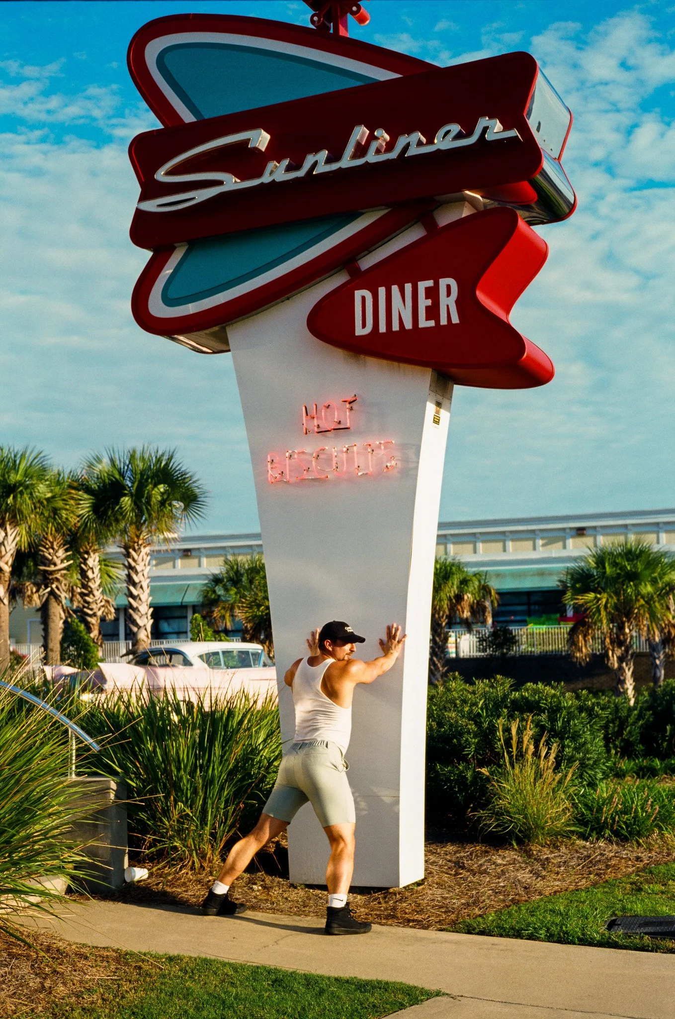 A person in a white tank top, beige shorts, and black boots posing in front of a vintage Sunliner diner sign with a neon "HOT & COLD" sign underneath, on a sunny day with palm trees in the background.